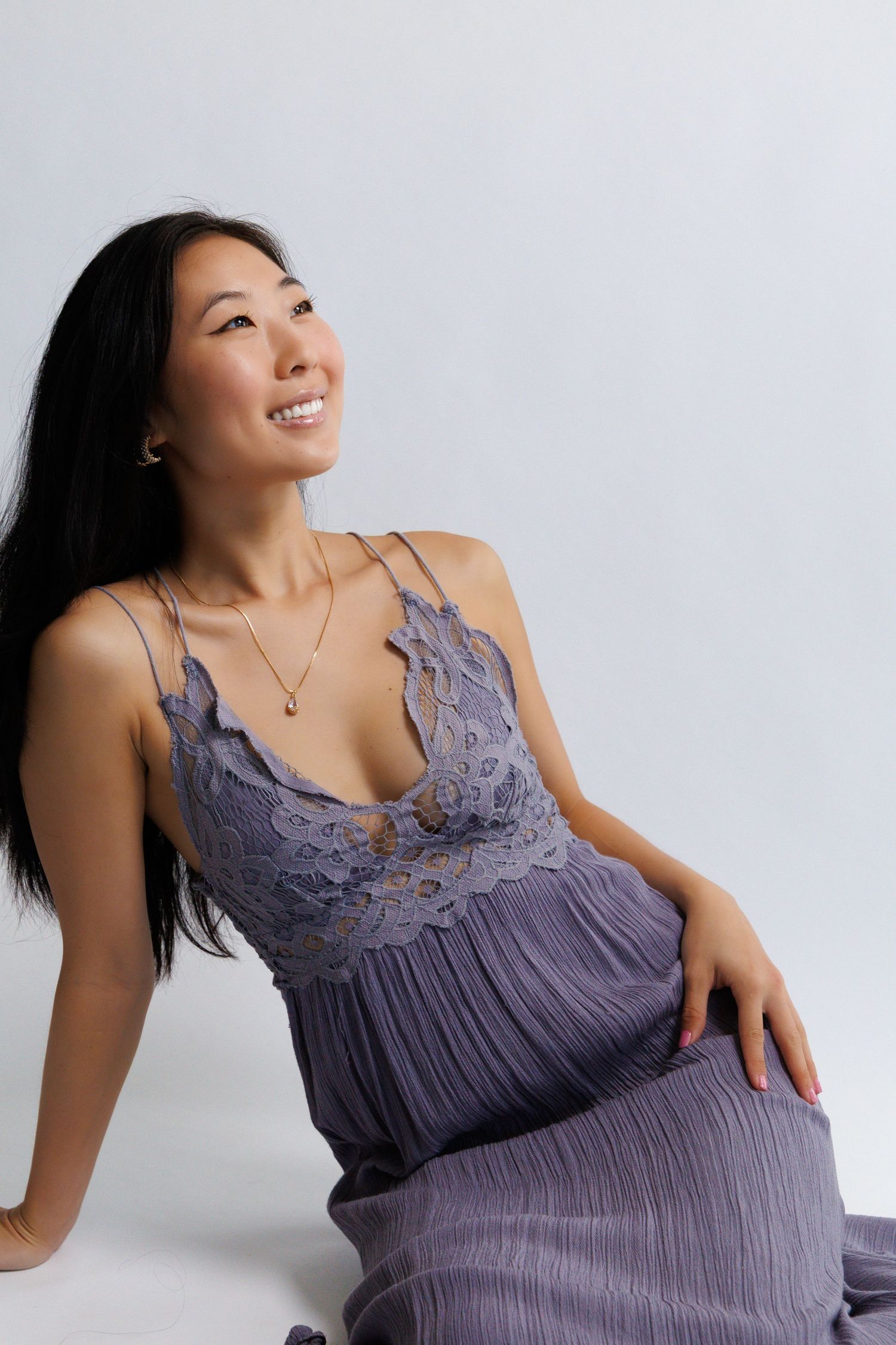 Smiling woman in a purple lace camisole dress sitting gracefully against a light gray background.