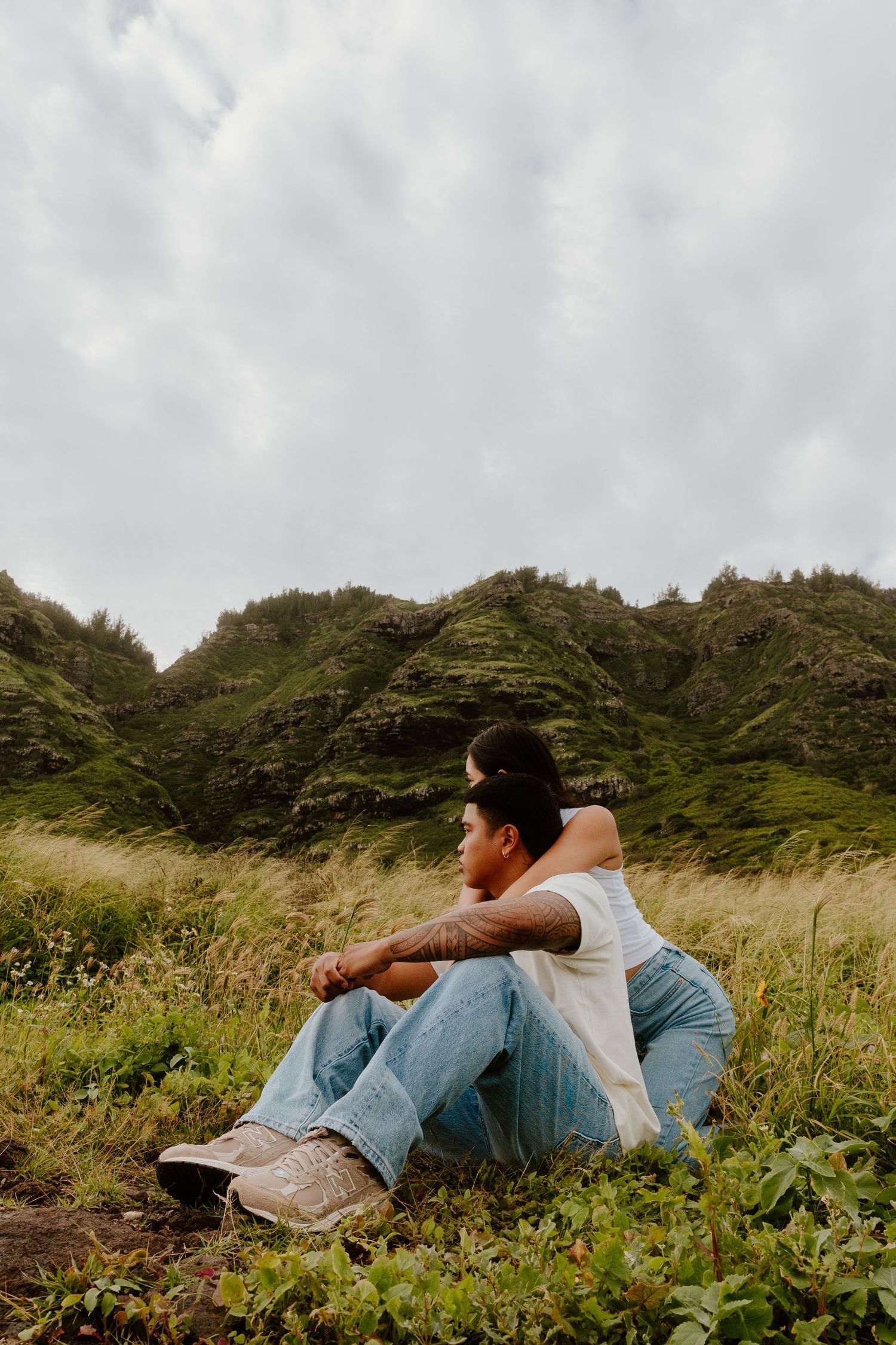 A young man with tattoos sits thoughtfully in a green meadow with dramatic misty mountains behind him.