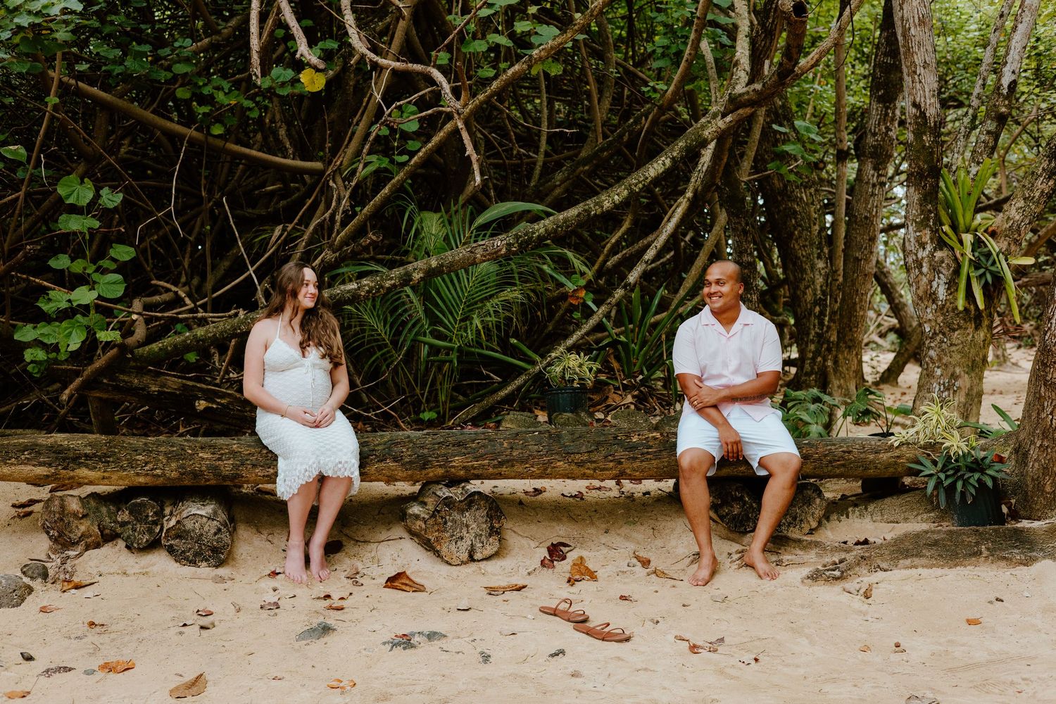 Pregnant couple sitting apart on a rustic bench on a sandy beach surrounded by tropical trees.
