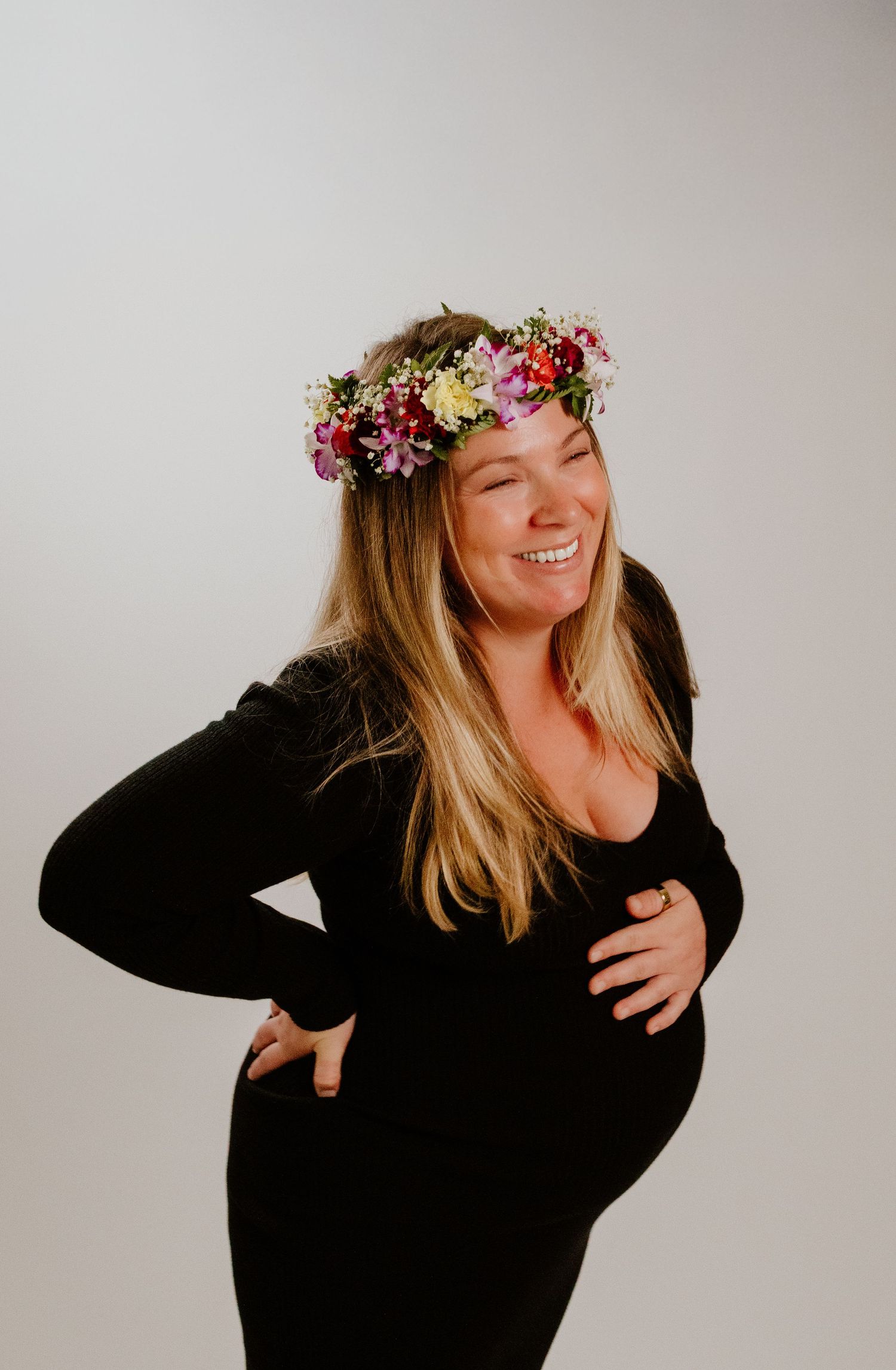 Laughing pregnant woman in black dress wearing floral crown during maternity photoshoot.