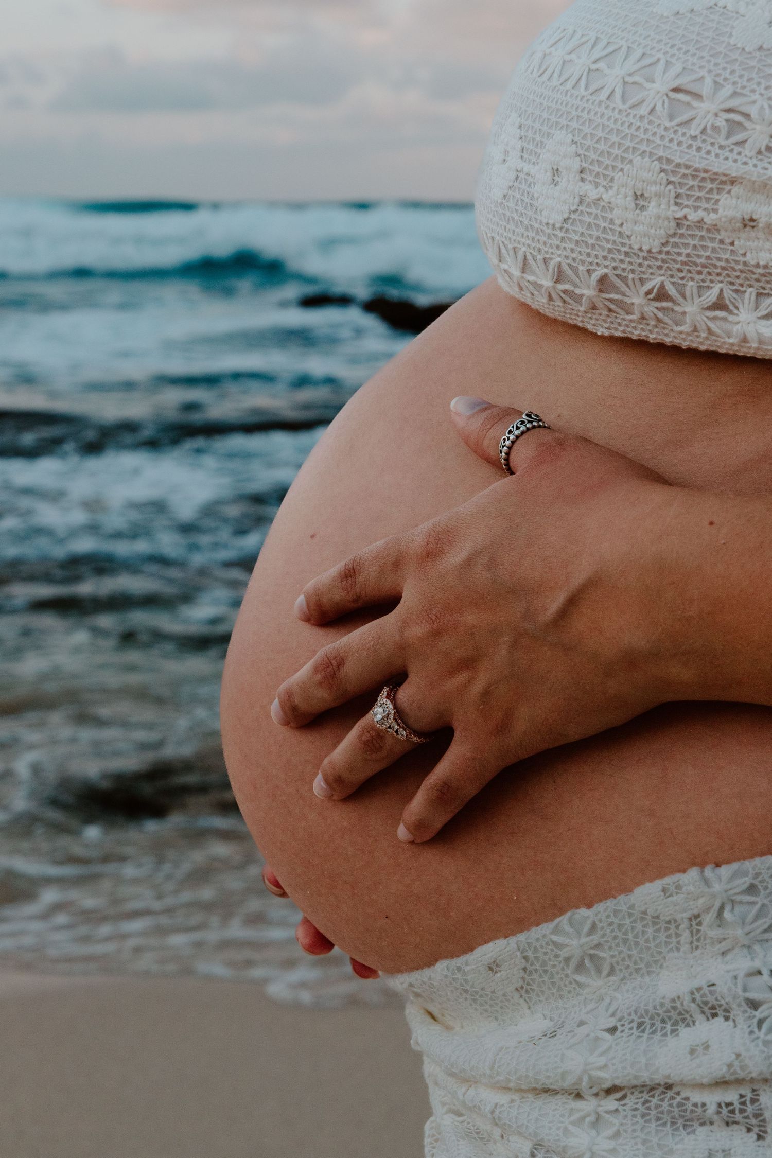 Close-up of pregnant belly with diamond rings and white lace dress at beach.