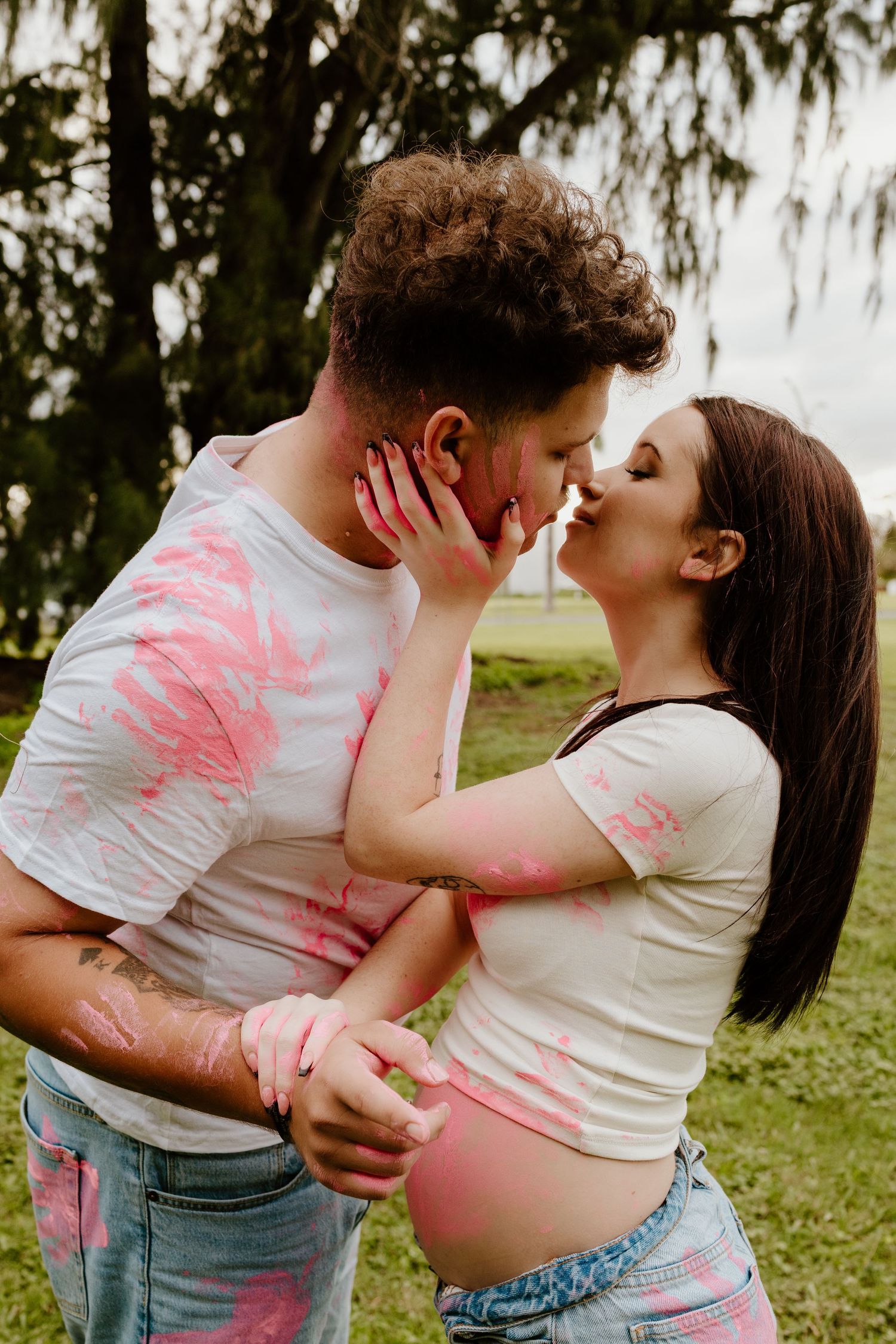 Expecting couple kissing outdoors covered in pink gender reveal paint in park setting.