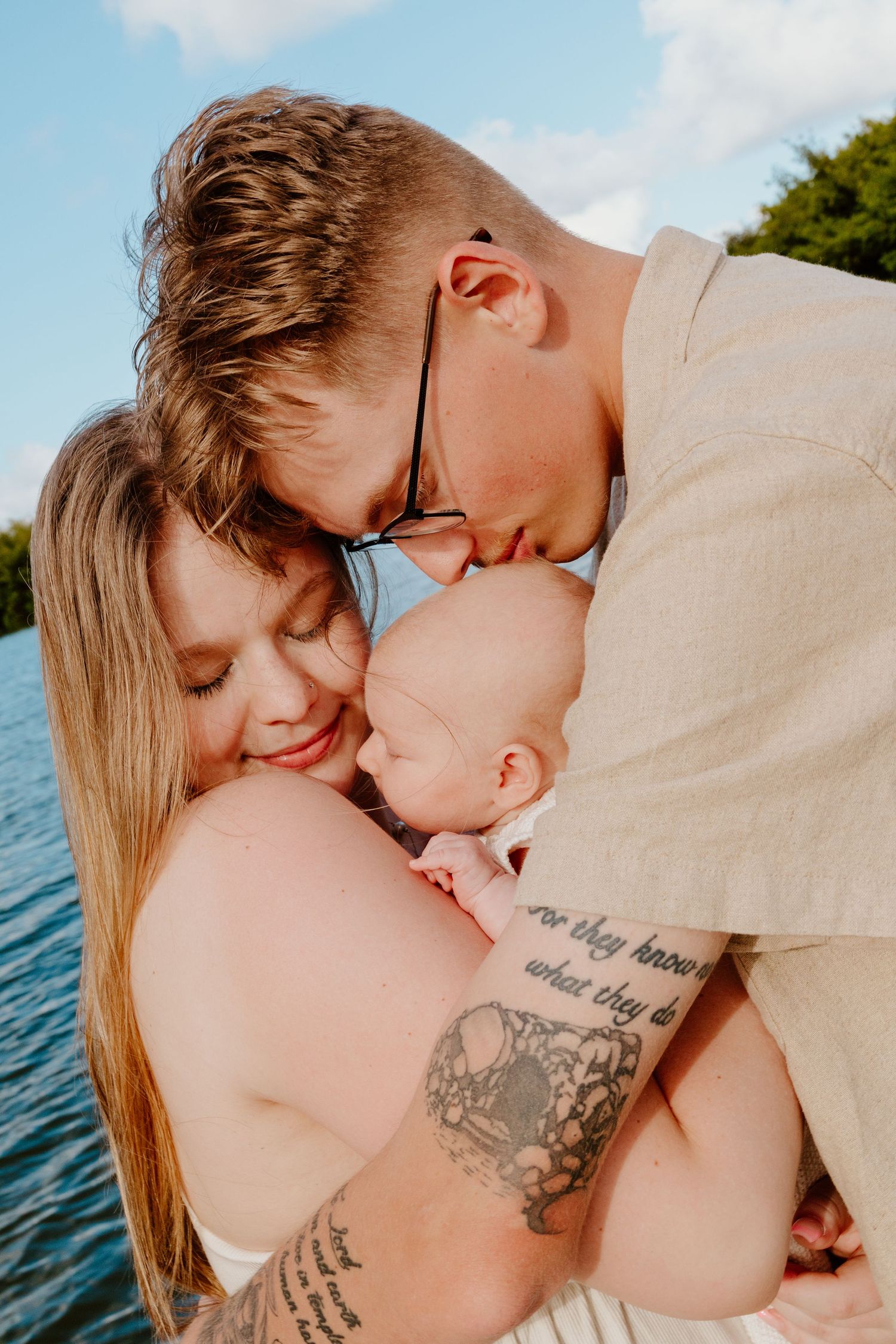 Tattooed parents kissing their newborn baby together near a sparkling blue lake.