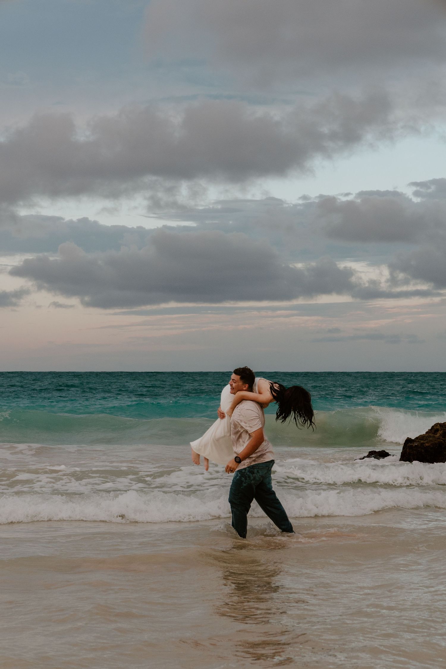 Man lifting woman in white dress while kissing on a tropical beach at sunset with turquoise waves.