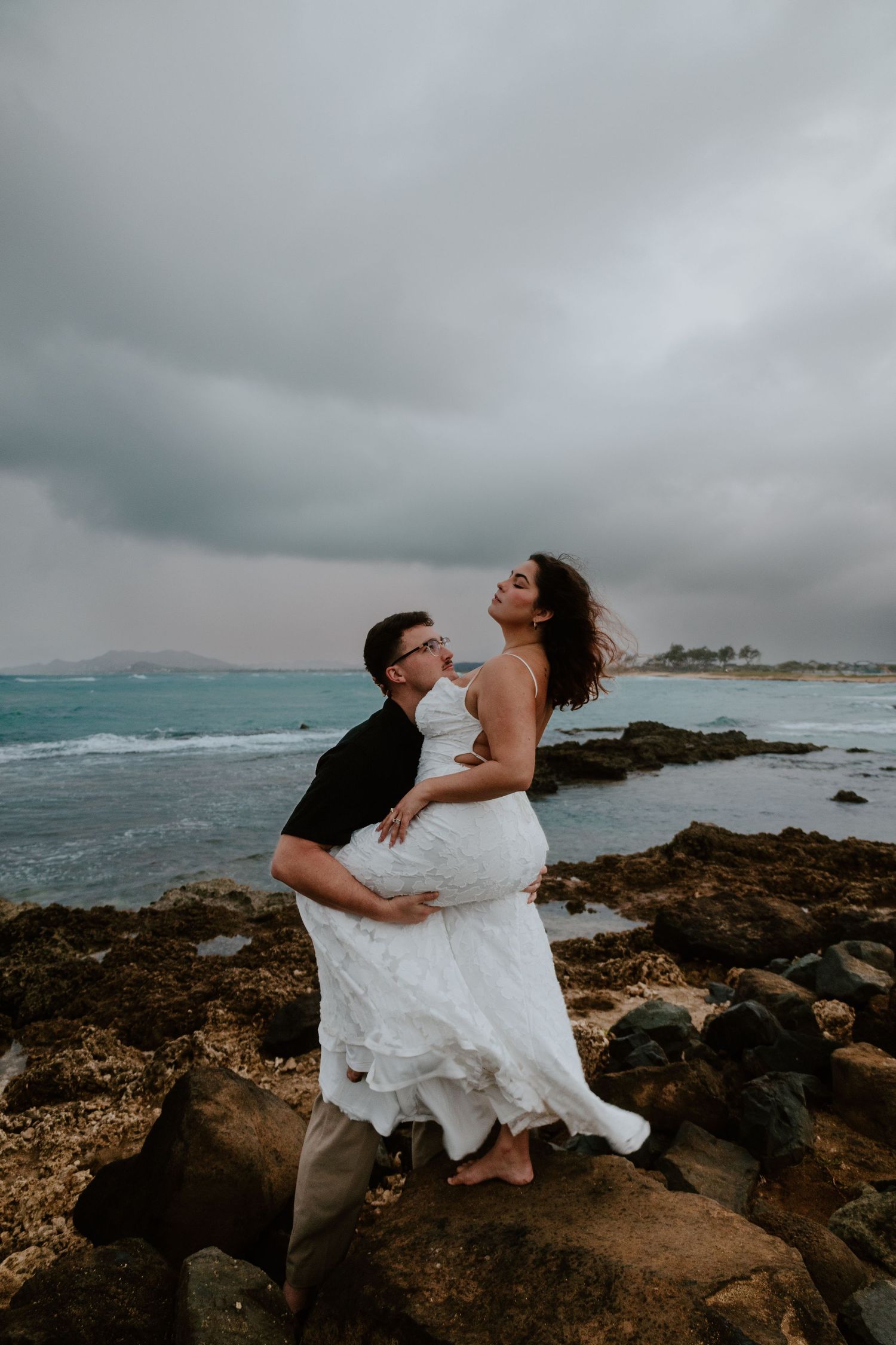 A groom in black shirt lifts his barefoot bride in white lace dress on rocky oceanside at dusk.
