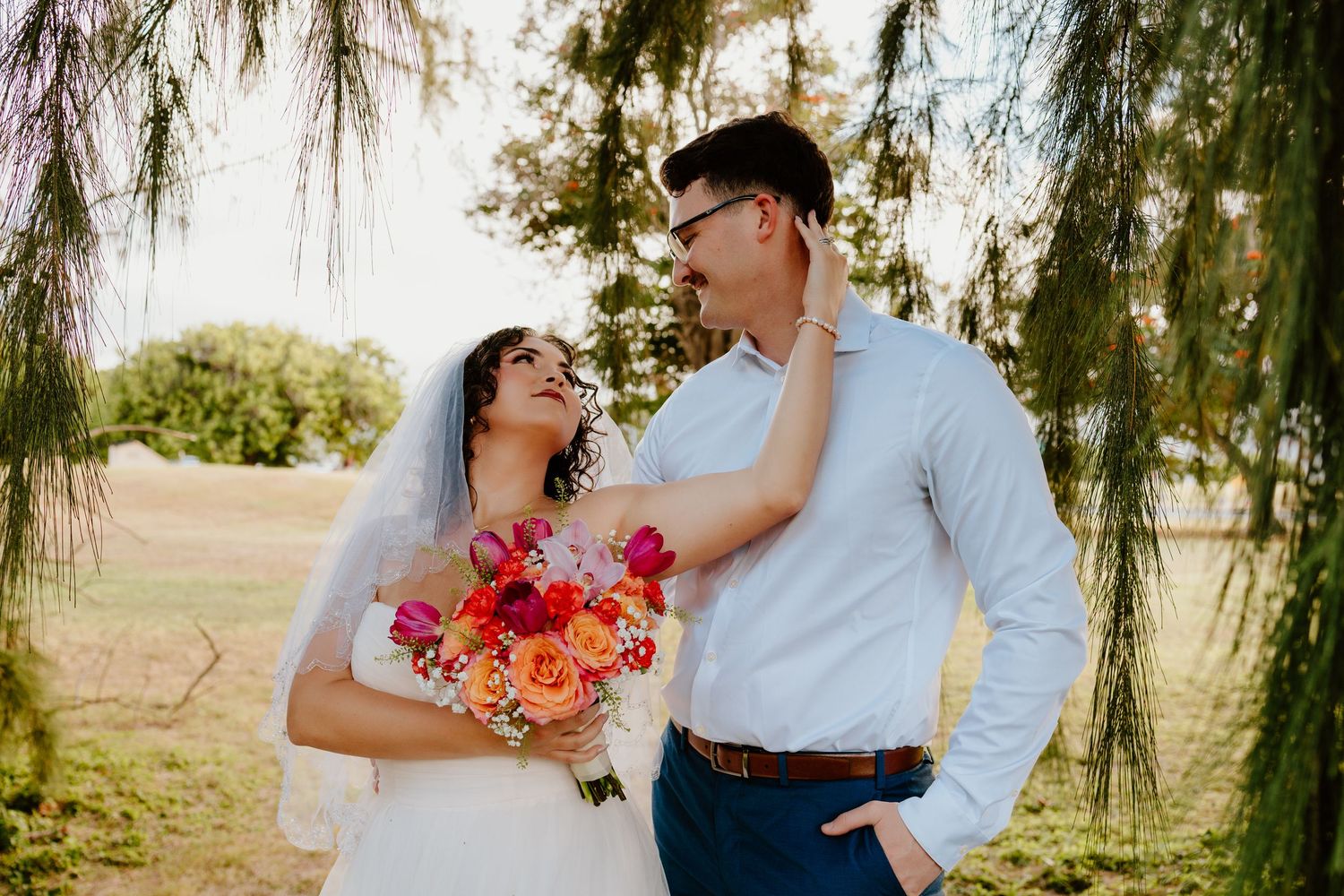 Bride and groom share a romantic moment under weeping willow, bride holding vibrant pink and orange bouquet.
