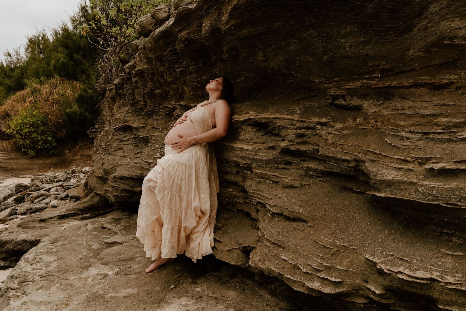 Pregnant woman in flowing beige dress leaning against layered rock formation outdoors.