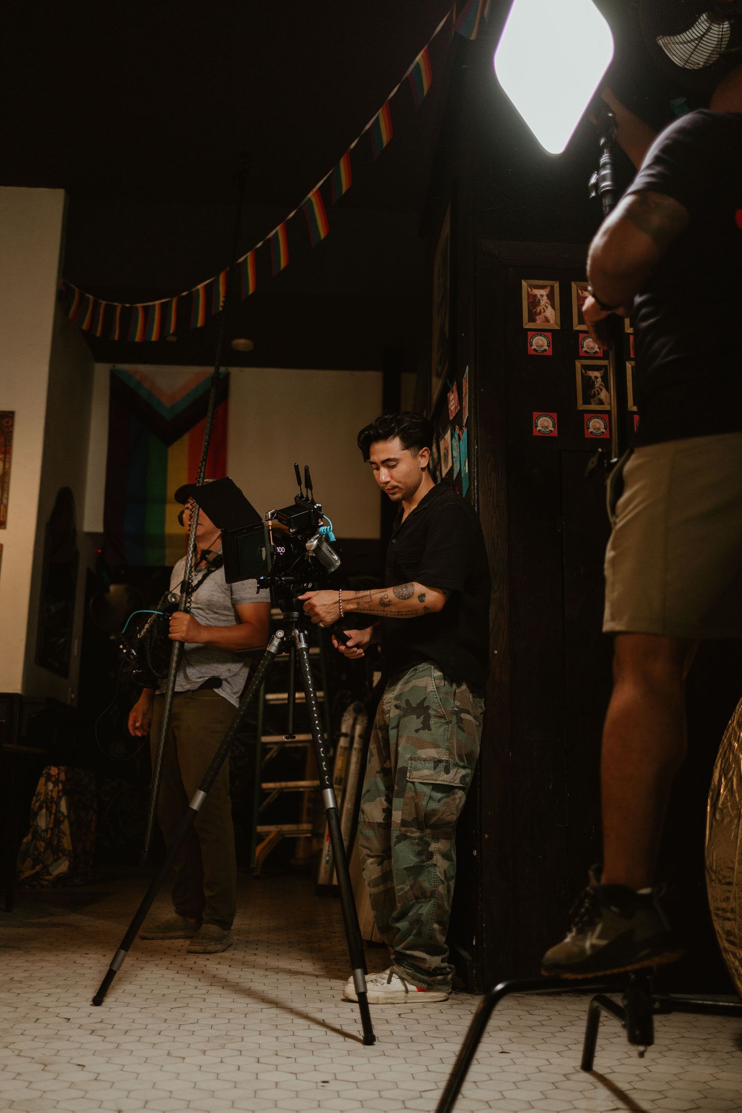 Film crew setting up a professional camera on a tripod inside a decorated room with rainbow flags and string lights.