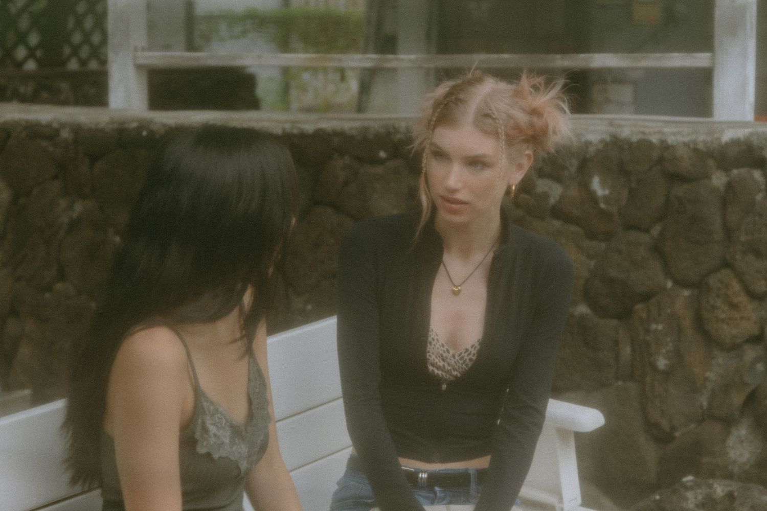 Two young women having a conversation while sitting outside against a stone wall.