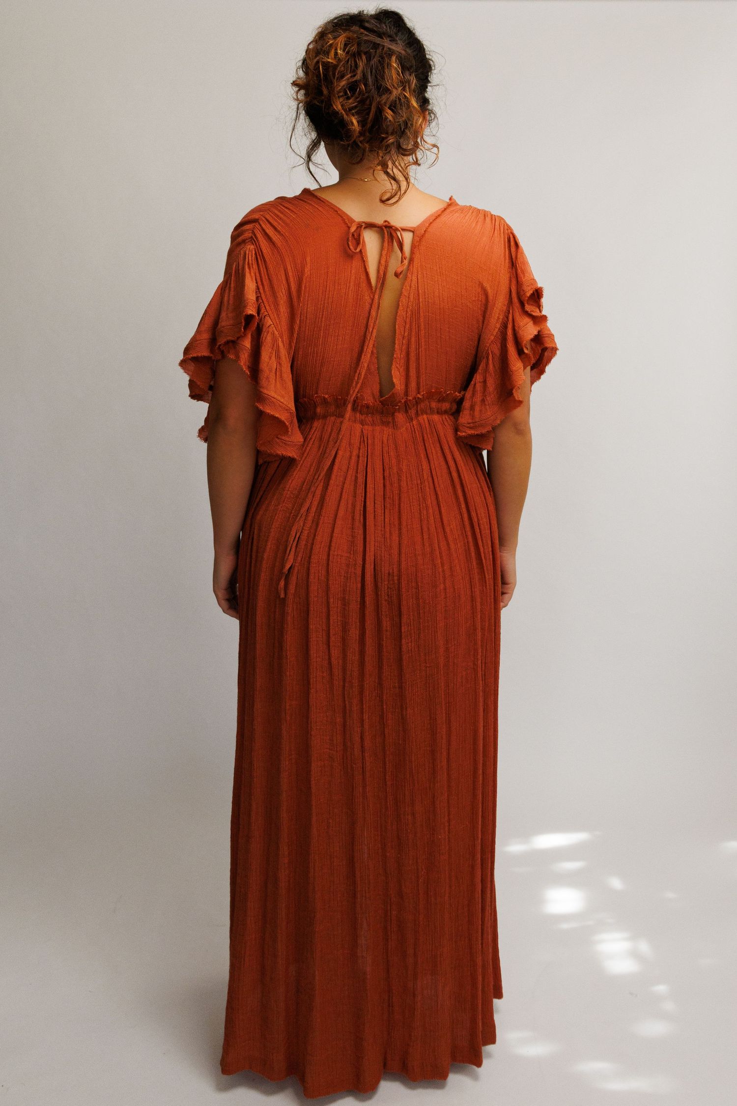 Back view of a woman wearing a rust orange maxi dress with flutter sleeves, cinched waist, and pleated skirt.