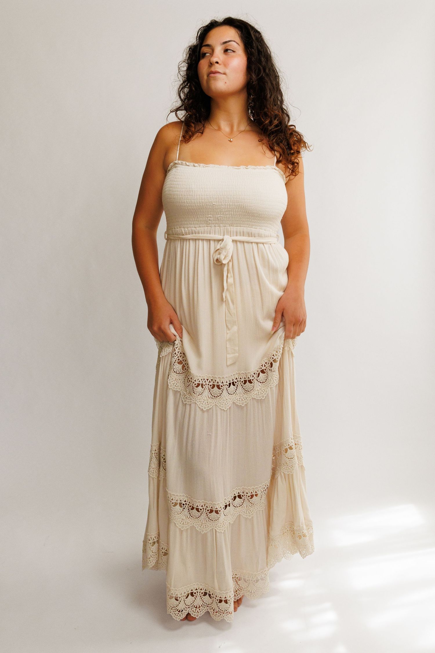 Woman wearing a cream boho maxi dress with crochet trim details and smocked bodice, spaghetti straps, tiered skirt.