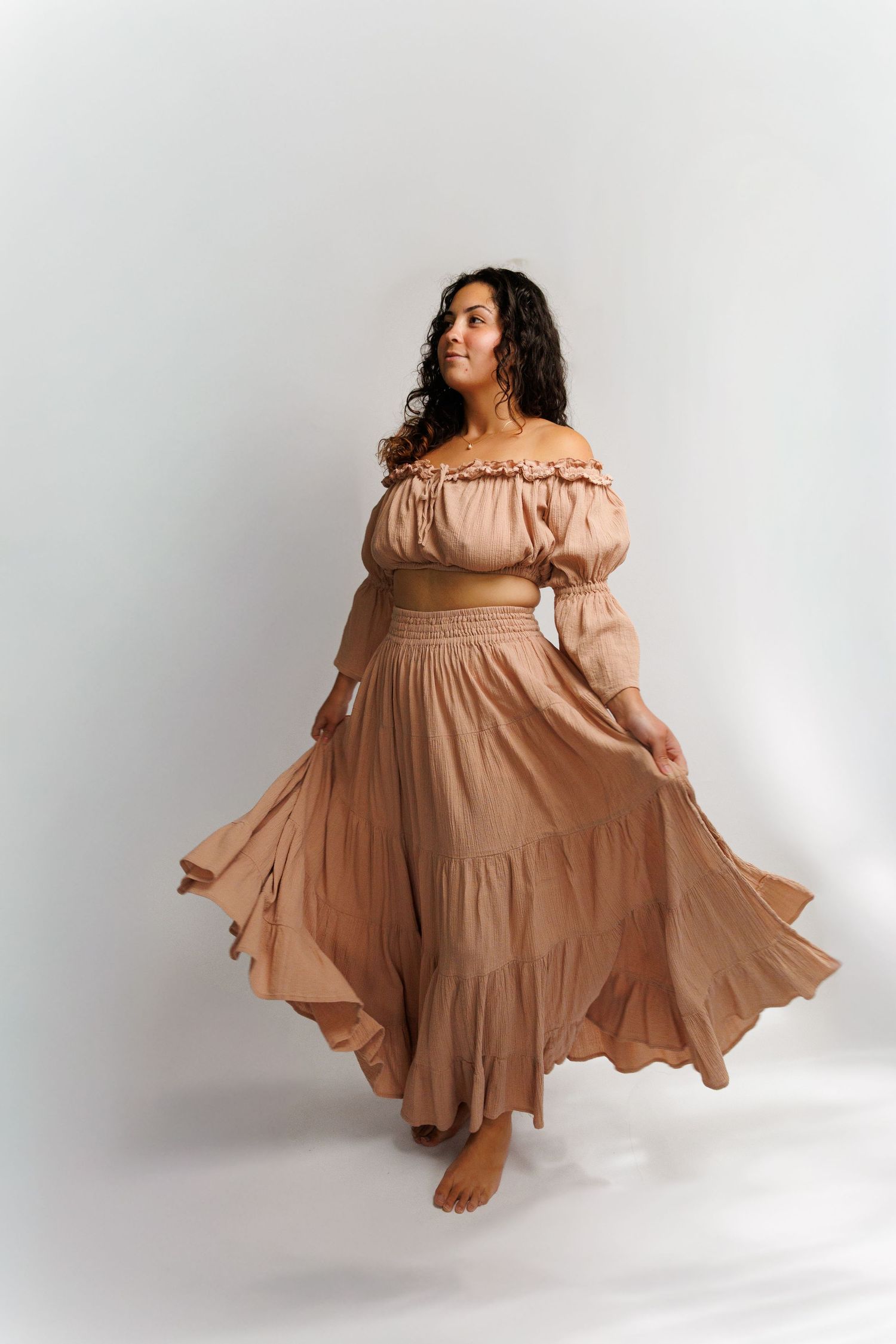 Woman in flowing beige off-shoulder crop top and maxi skirt set, twirling gracefully against white background.