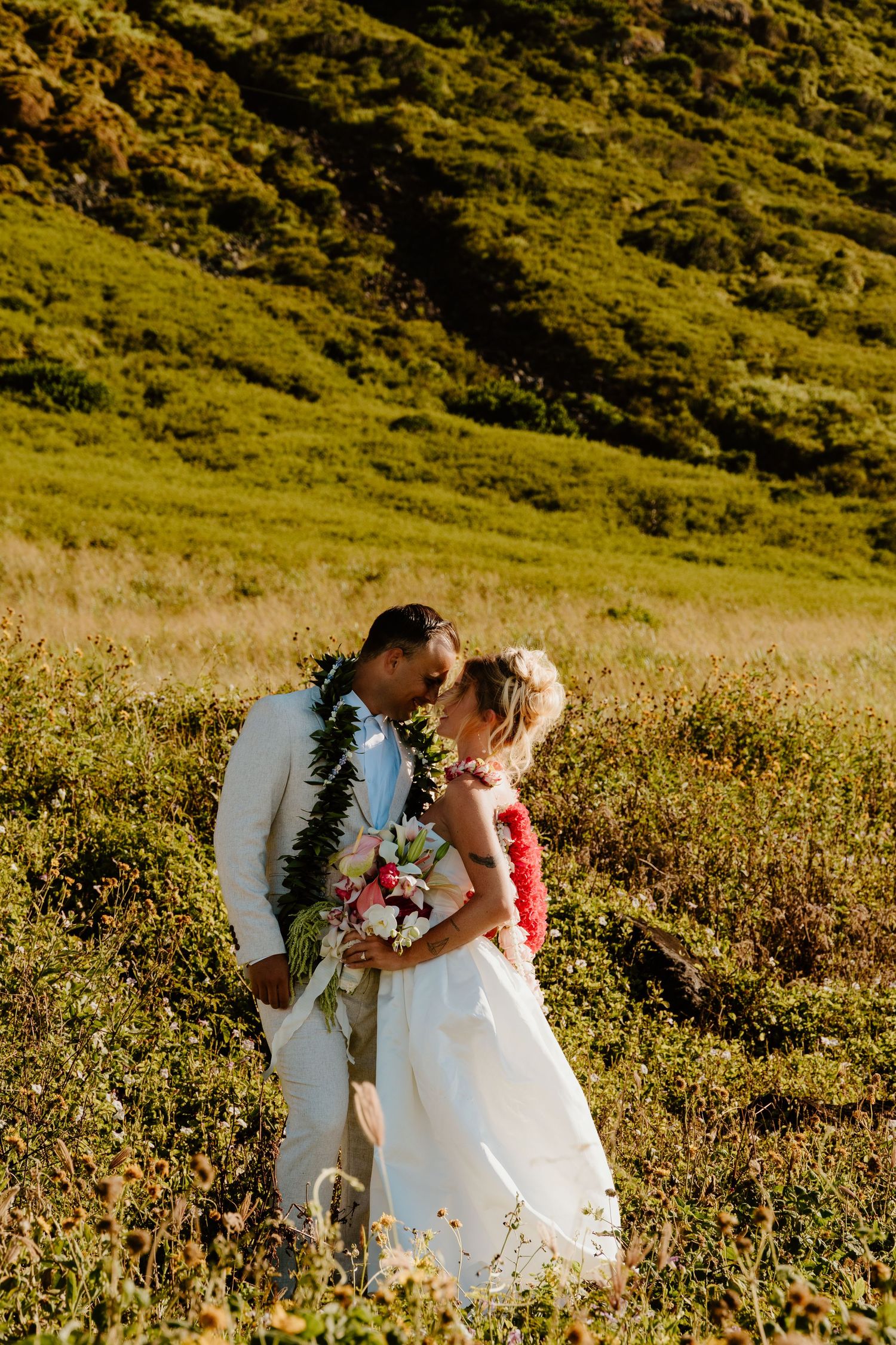 Newlyweds sharing tender kiss wearing floral leis in golden sunlit Hawaiian hillside meadow.