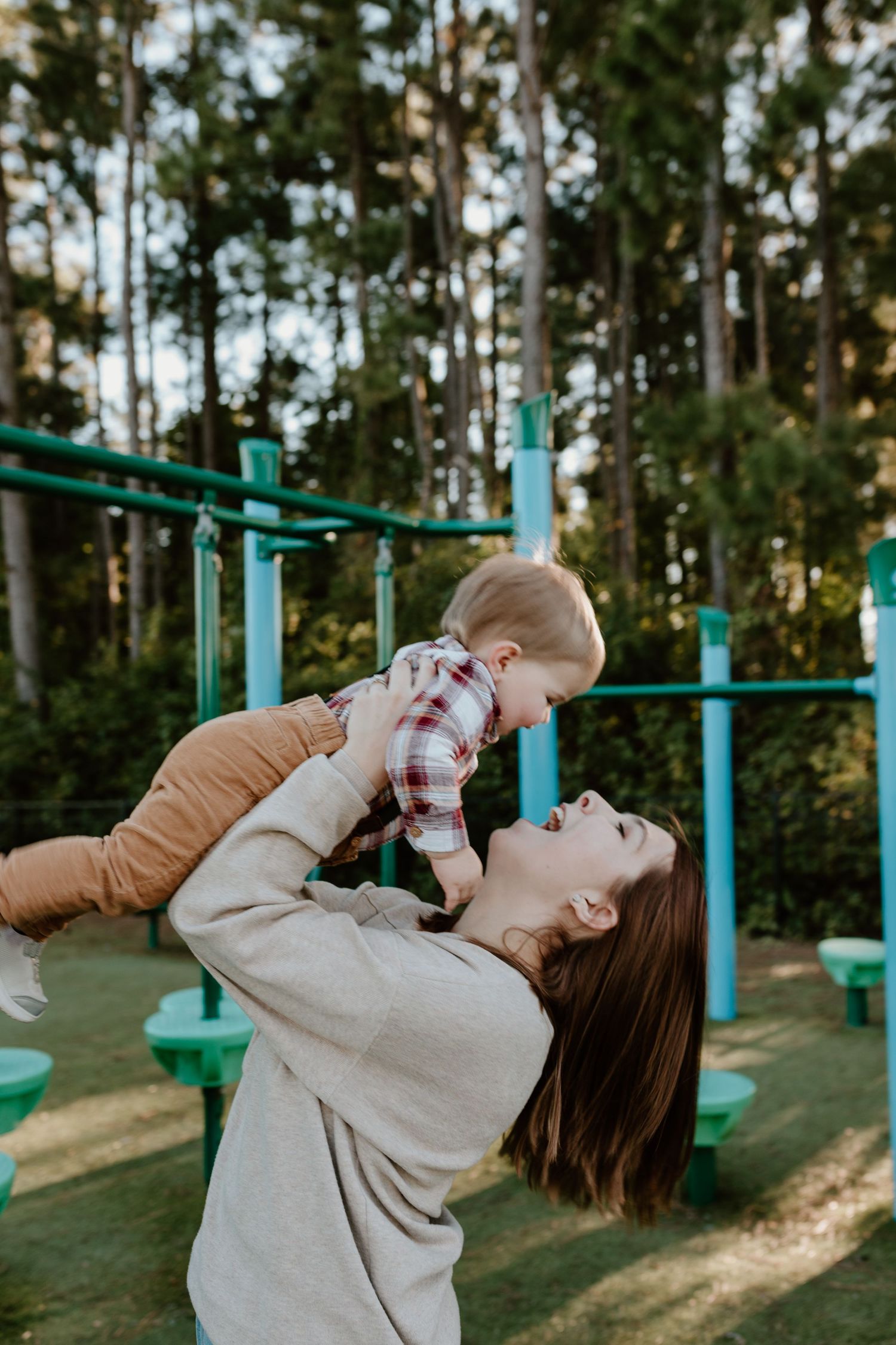 Smiling mother in beige sweater lifts laughing toddler in plaid shirt high in the air near a playground.