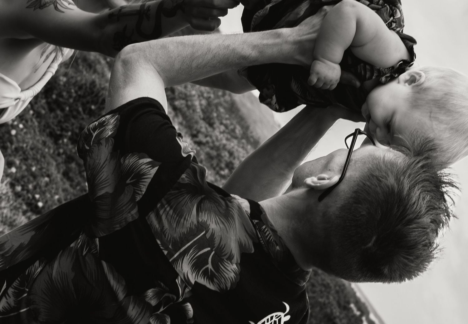 Black and white photo of father lifting smiling baby while mother watches outdoors.