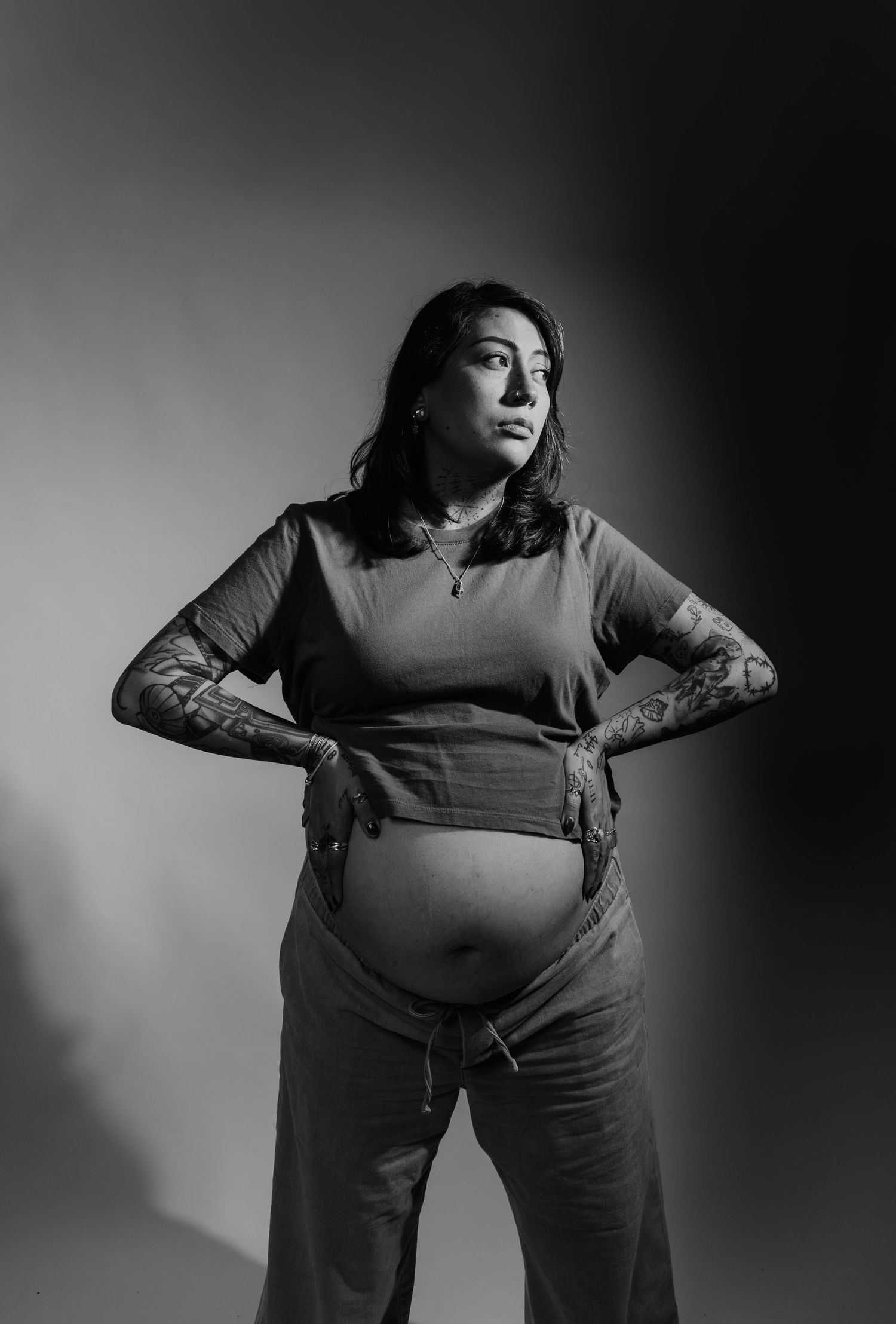 Black and white portrait of pregnant tattooed woman posing with hands on hips.