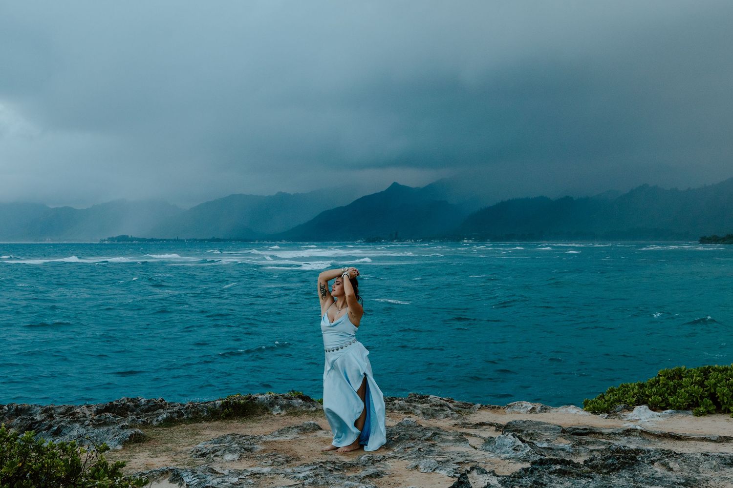 Woman in blue dress standing on rocky shore with turquoise ocean and dramatic stormy mountains in background.