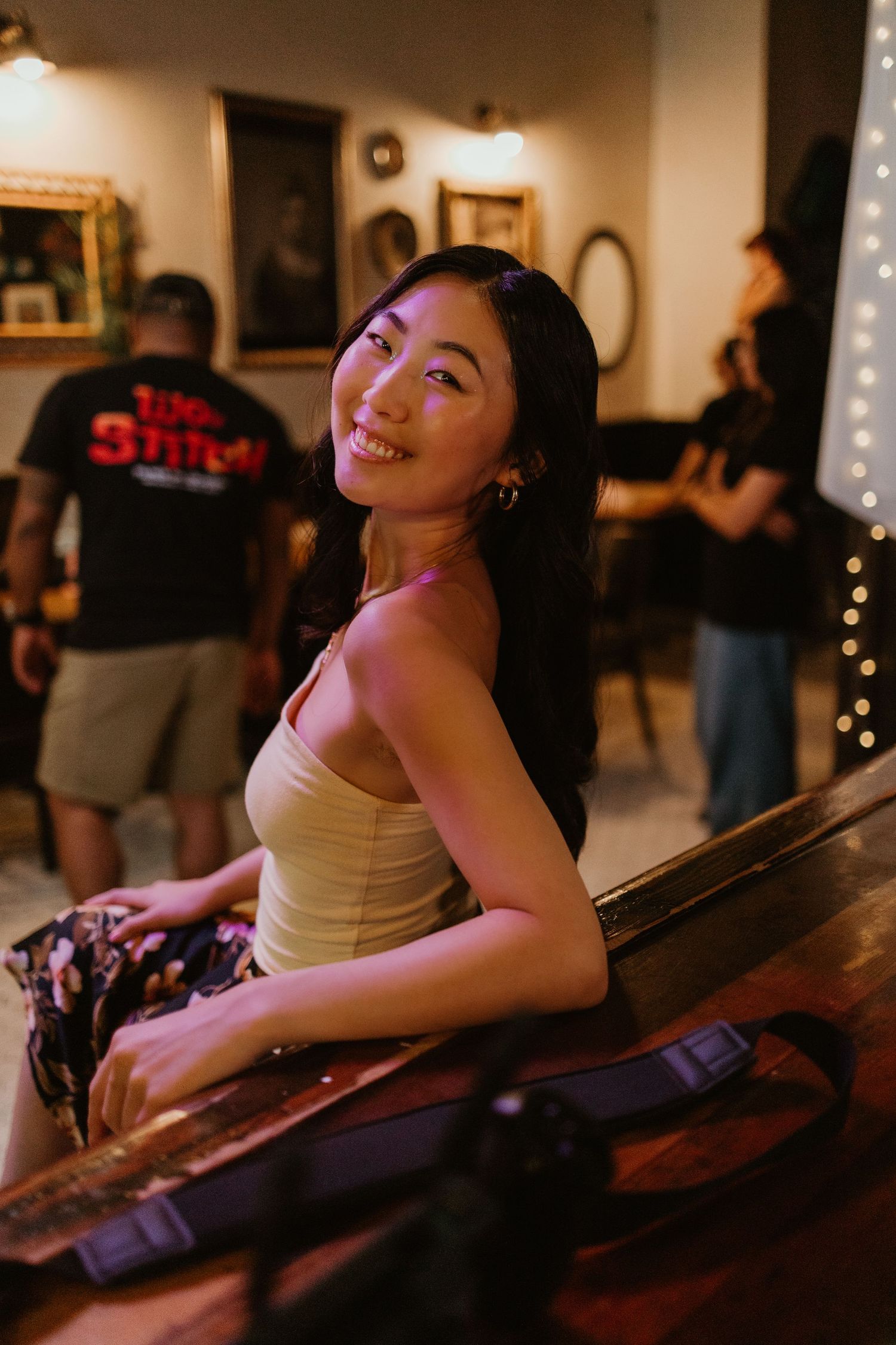 Smiling Asian woman leaning on a wooden bar counter in a warmly lit restaurant setting.