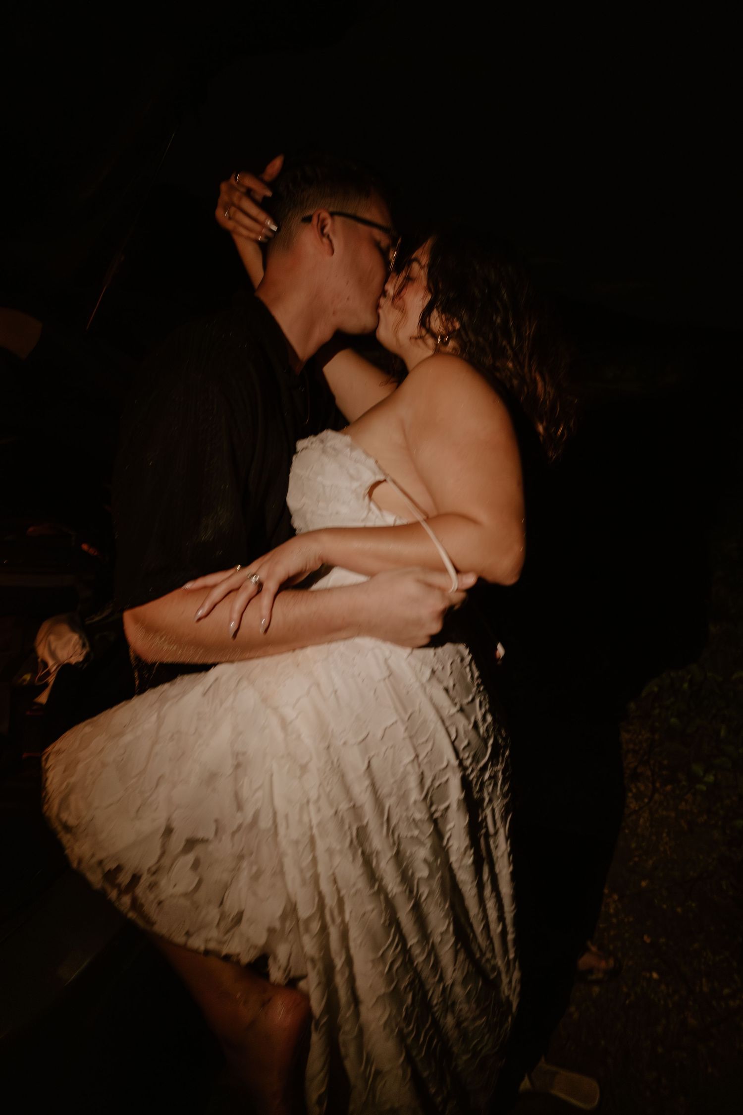 A bride and groom share a romantic kiss at night, the bride in a short white lace dress.
