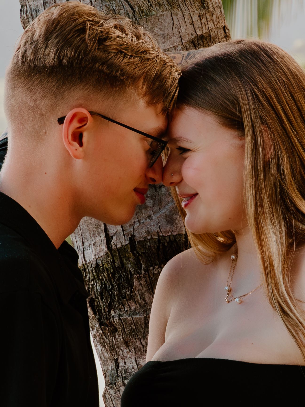 A young couple shares a romantic forehead-to-forehead moment leaning against a palm tree outdoors.
