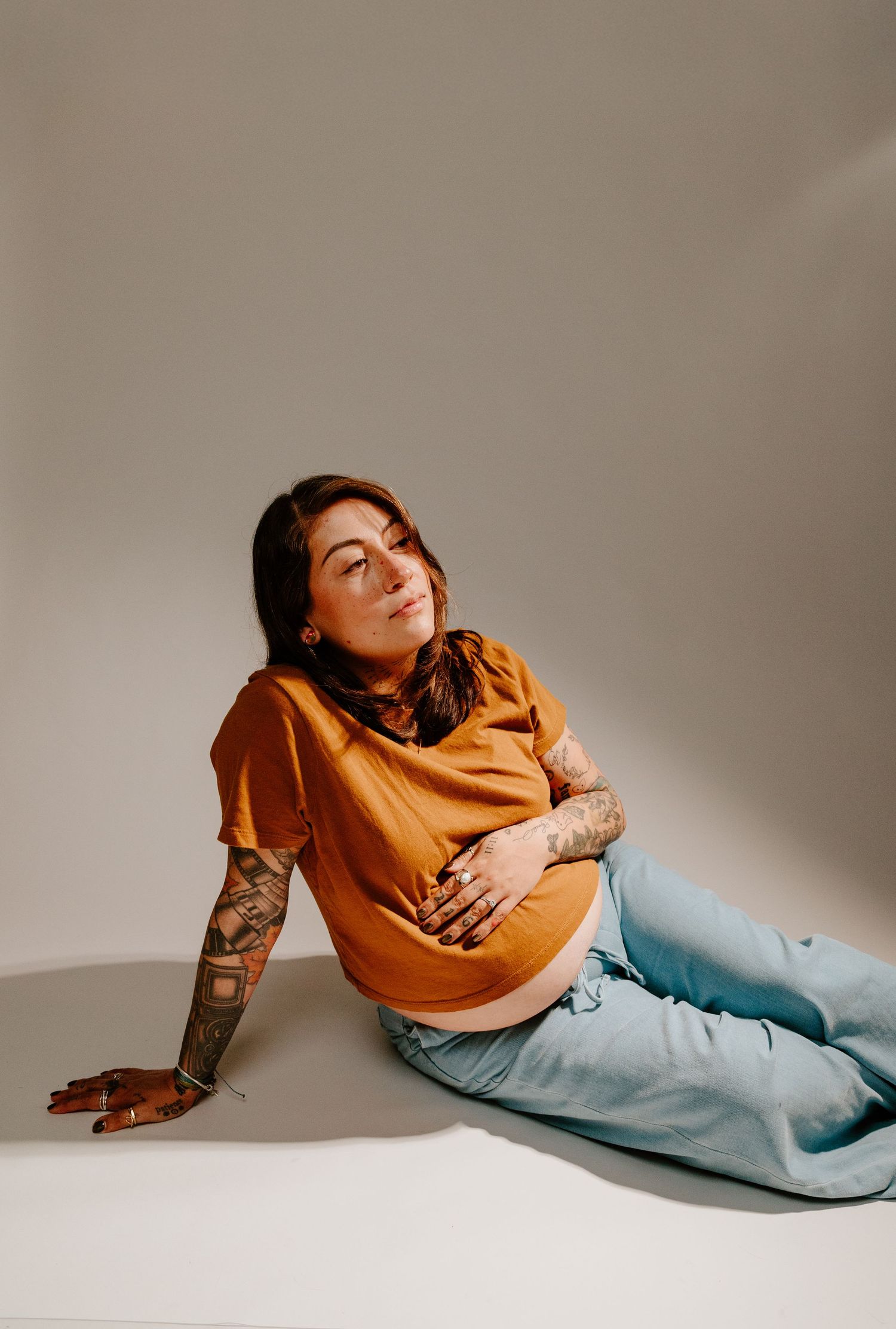 Tattooed pregnant woman in orange tee and jeans sitting on floor, cradling bump, looking up.