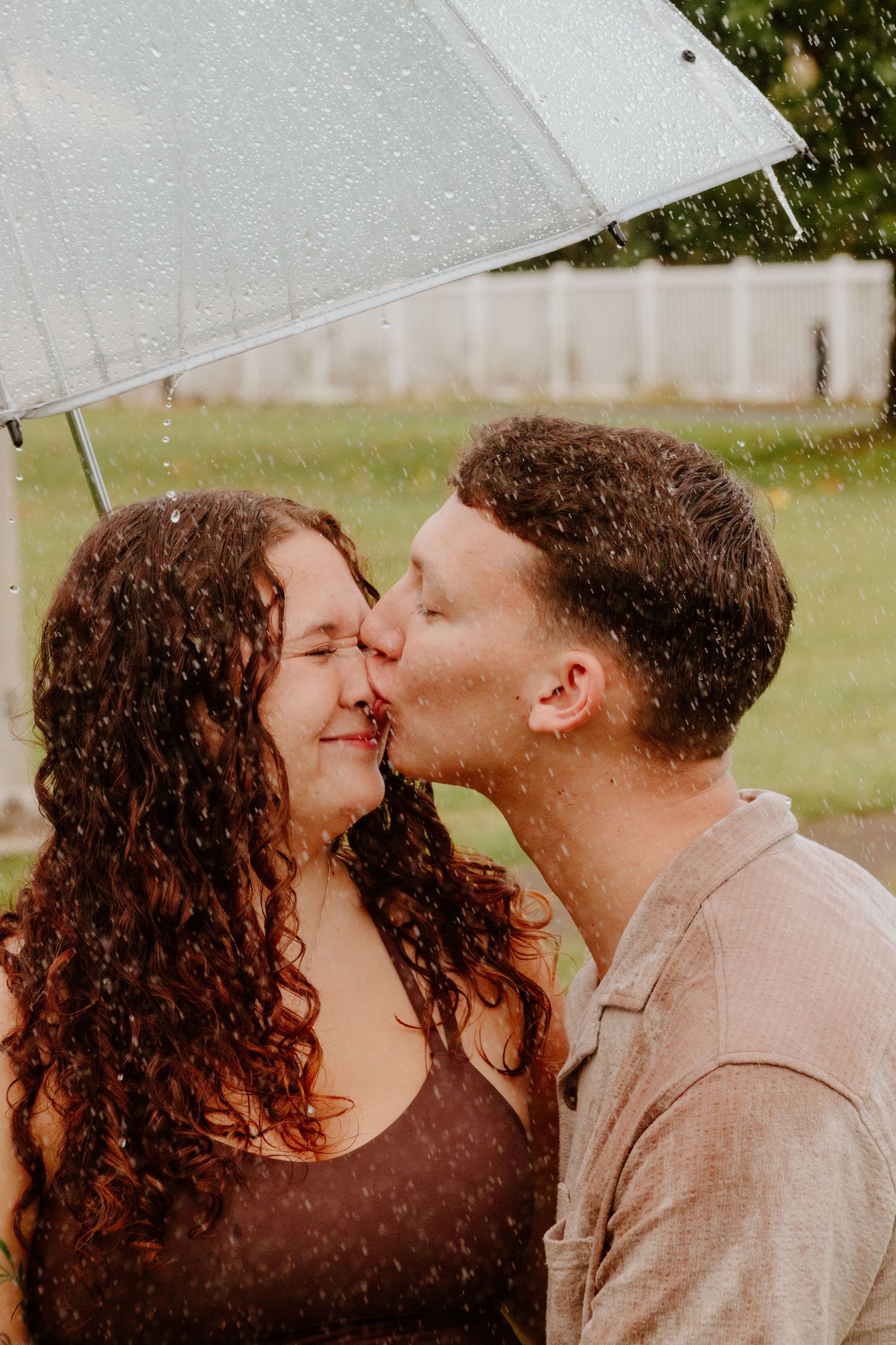 Couple kissing under a clear umbrella in the rain outdoors.