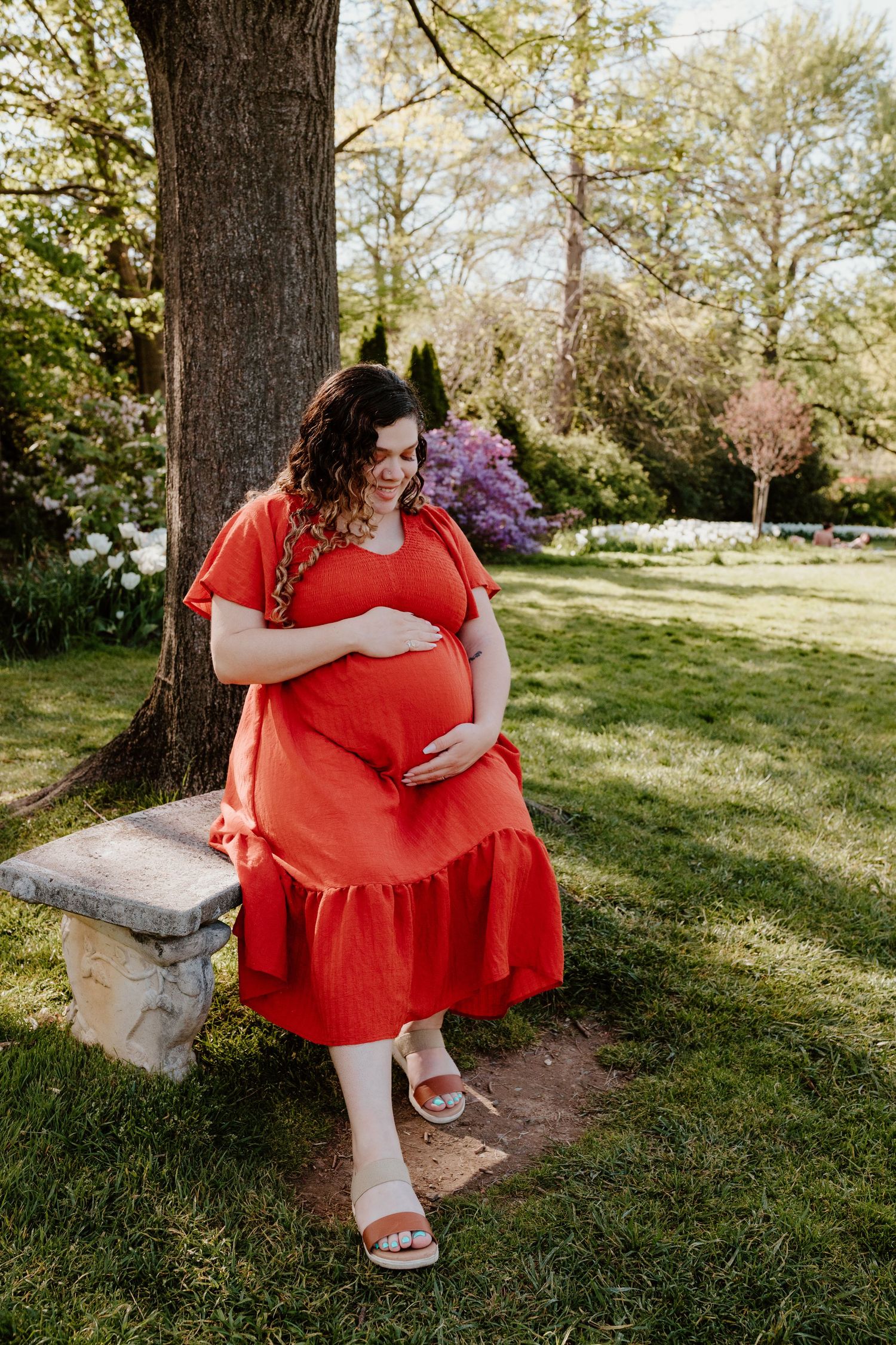 Pregnant woman in red dress cradling baby bump while leaning against a tree in a blooming spring garden.