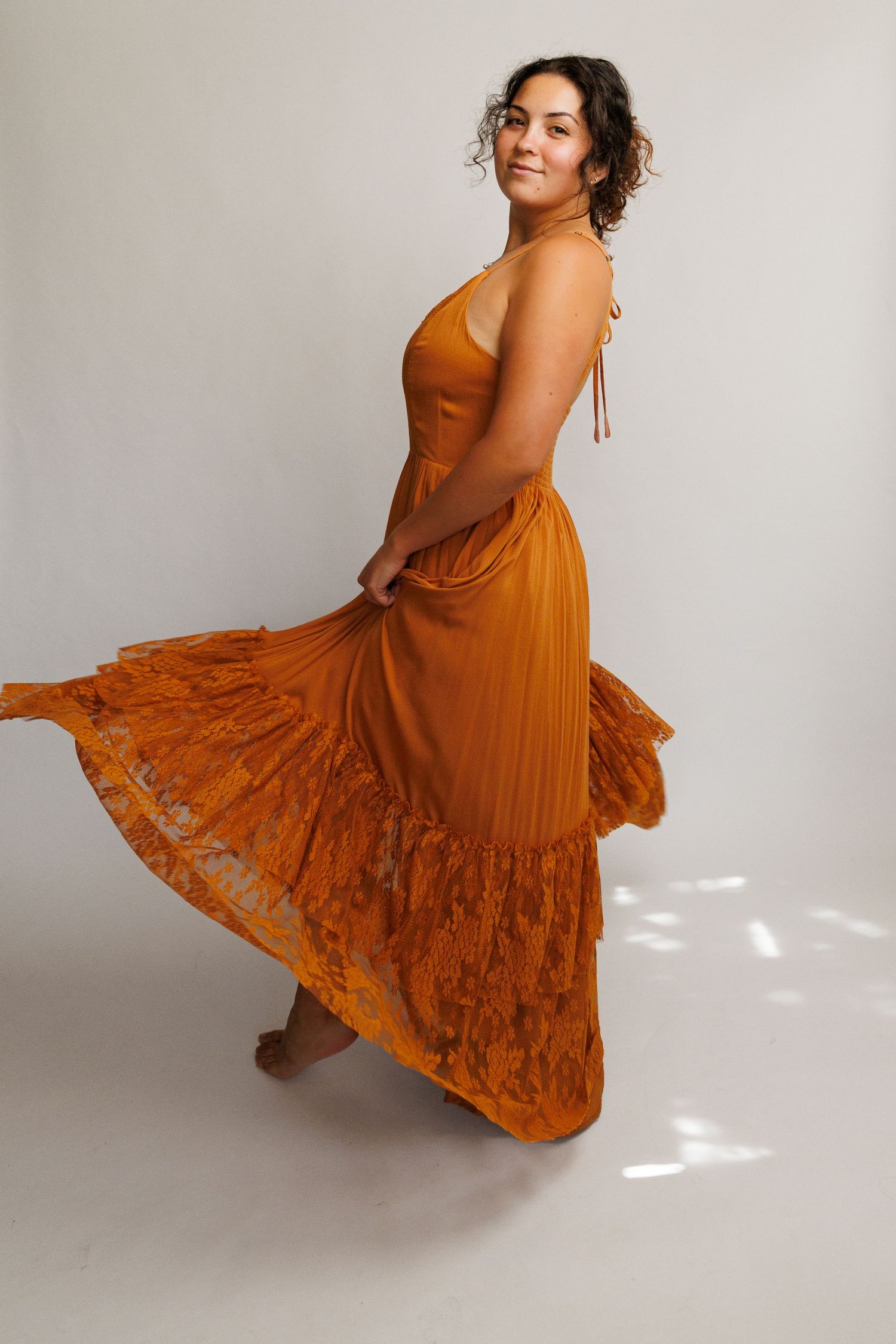 Woman twirling in a flowing burnt orange halter maxi dress with lace hem details on a white background.