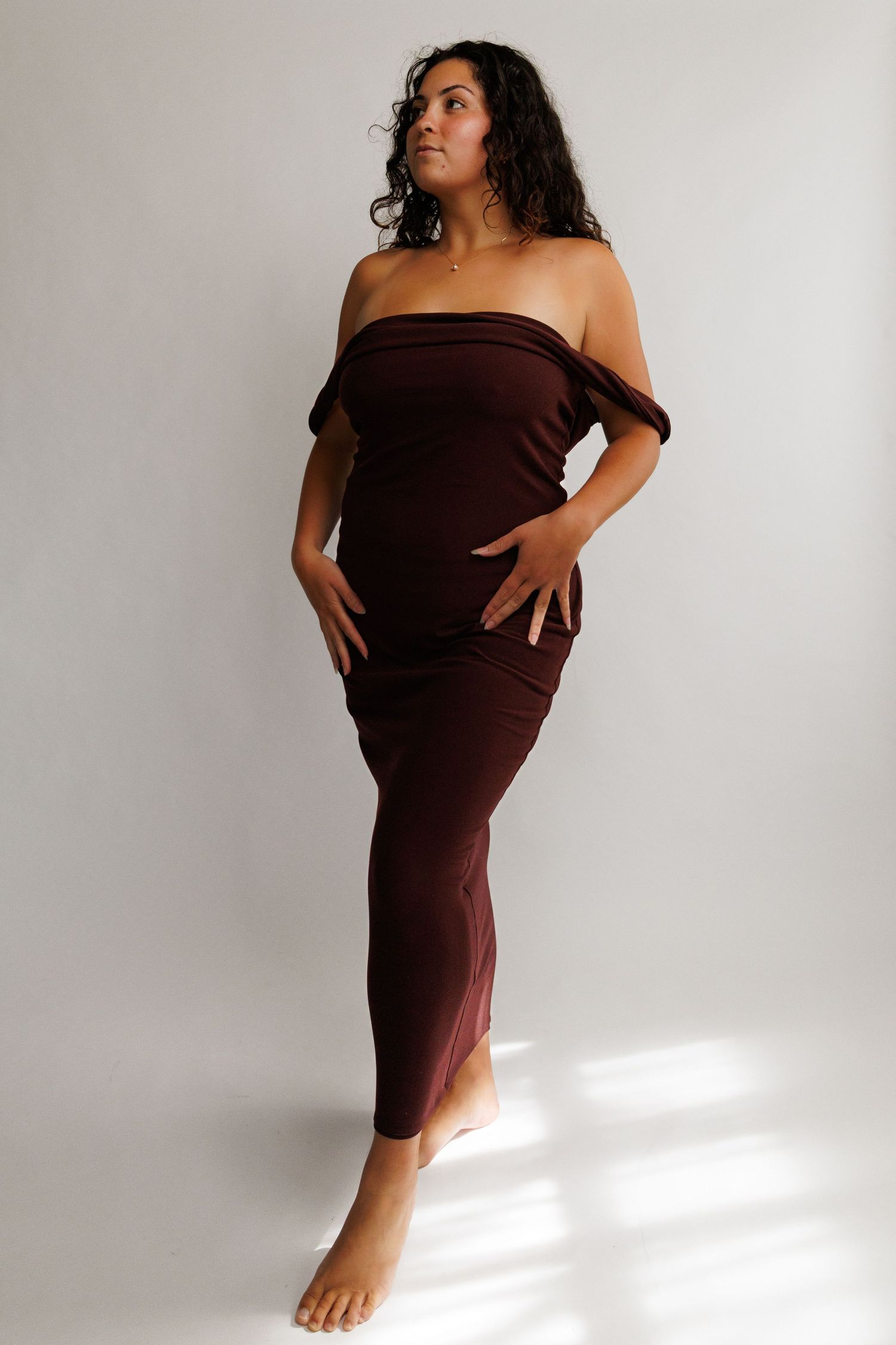 Woman wearing a dark brown off-shoulder maxi dress, posing barefoot against a light gray background.