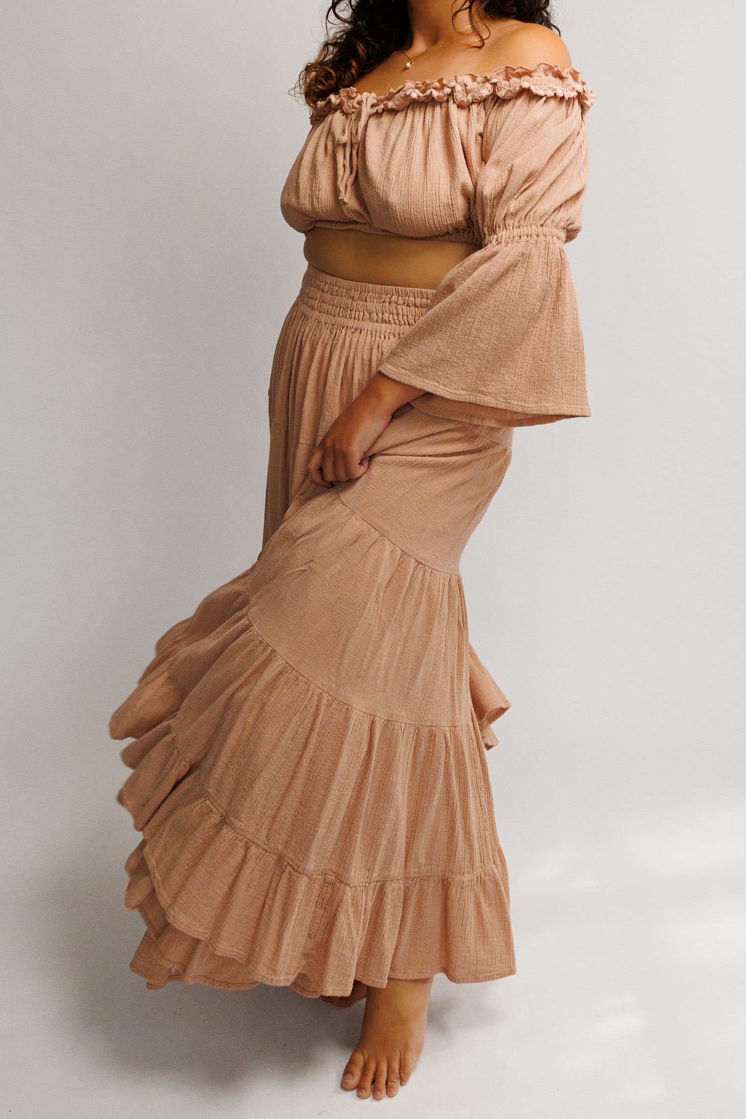 Woman wearing a blush pink off-shoulder crop top and matching tiered maxi skirt set with ruffle details.