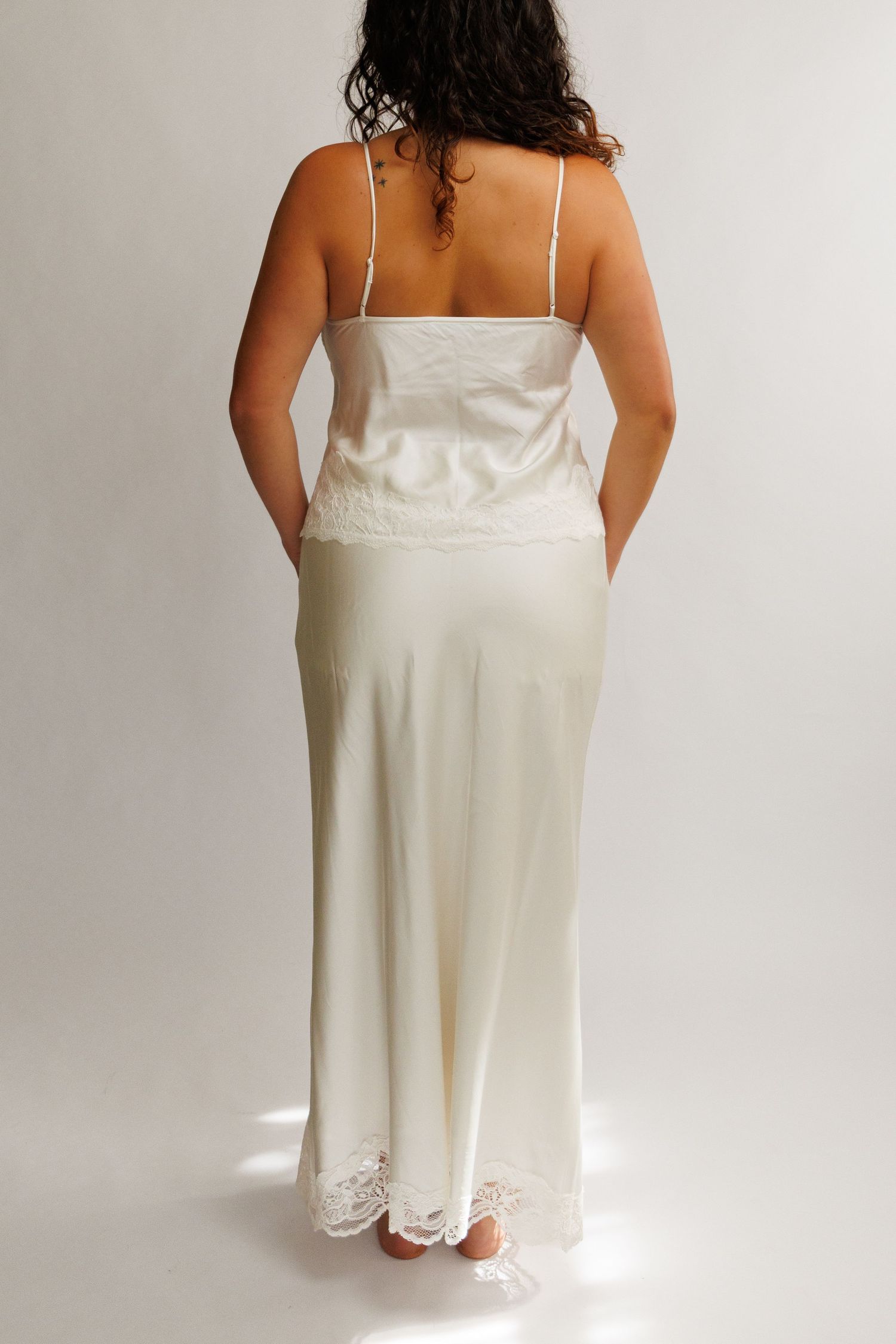 Woman wearing ivory satin slip dress with lace trim and spaghetti straps, shown from behind on white background.