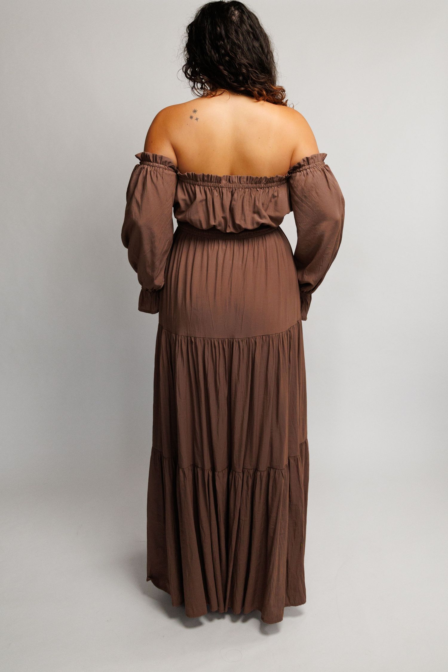 Back view of a woman wearing a brown off-shoulder tiered maxi dress with long sleeves and ruffle details.