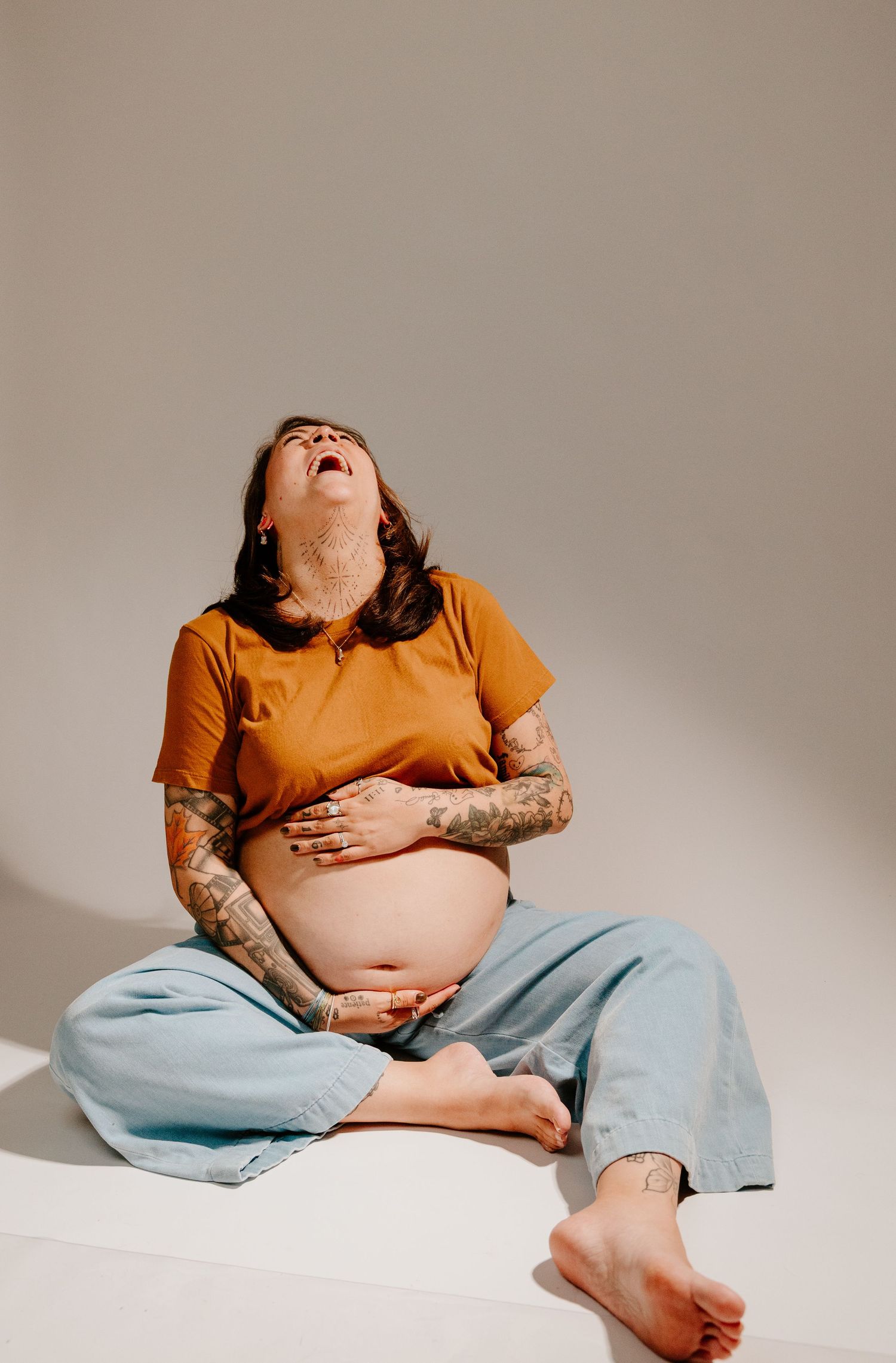 Tattooed pregnant woman in orange shirt sitting barefoot, head back laughing joyfully.