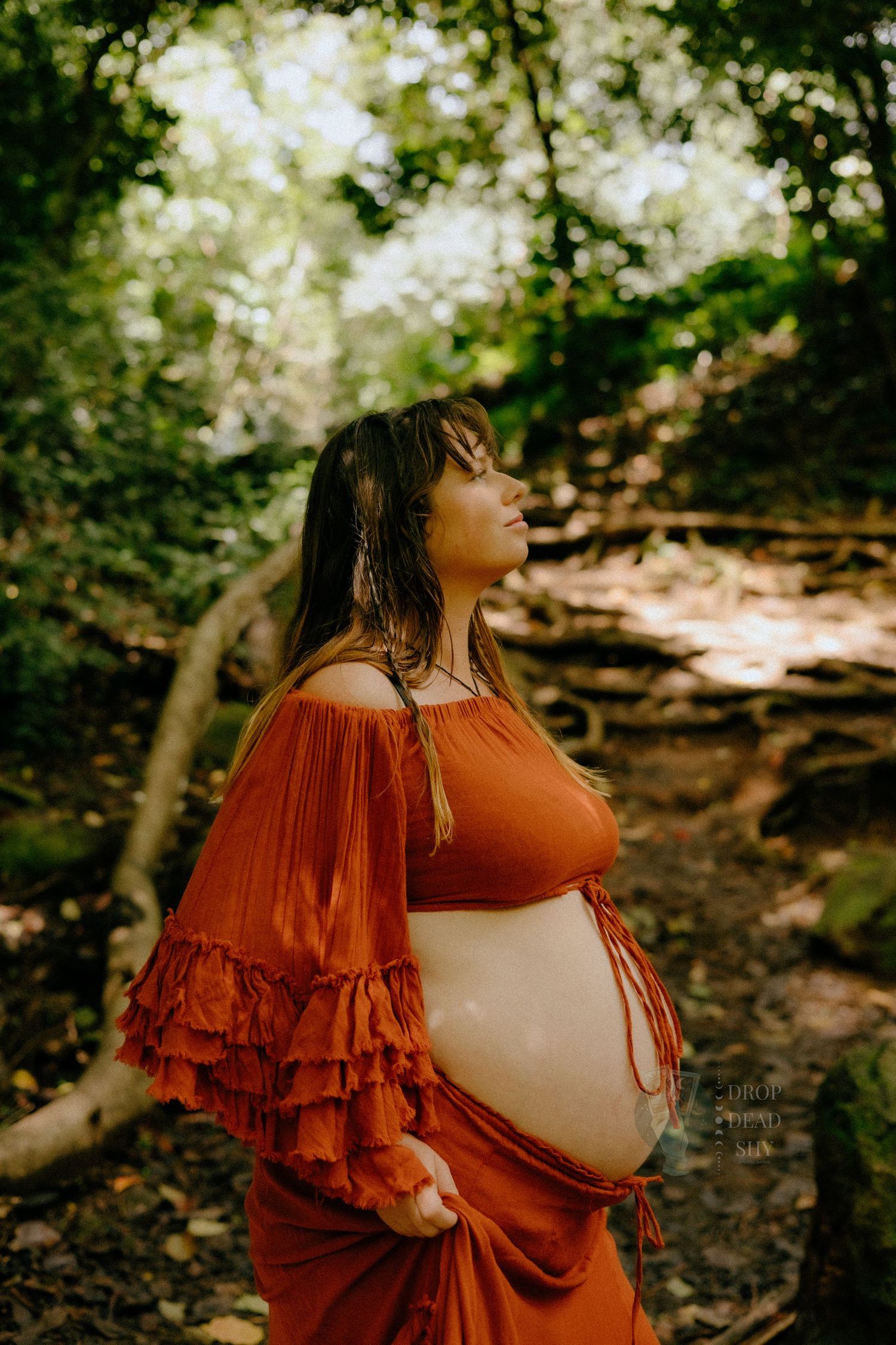 Beautiful pregnant woman in orange boho dress gazing upward in serene forest.
