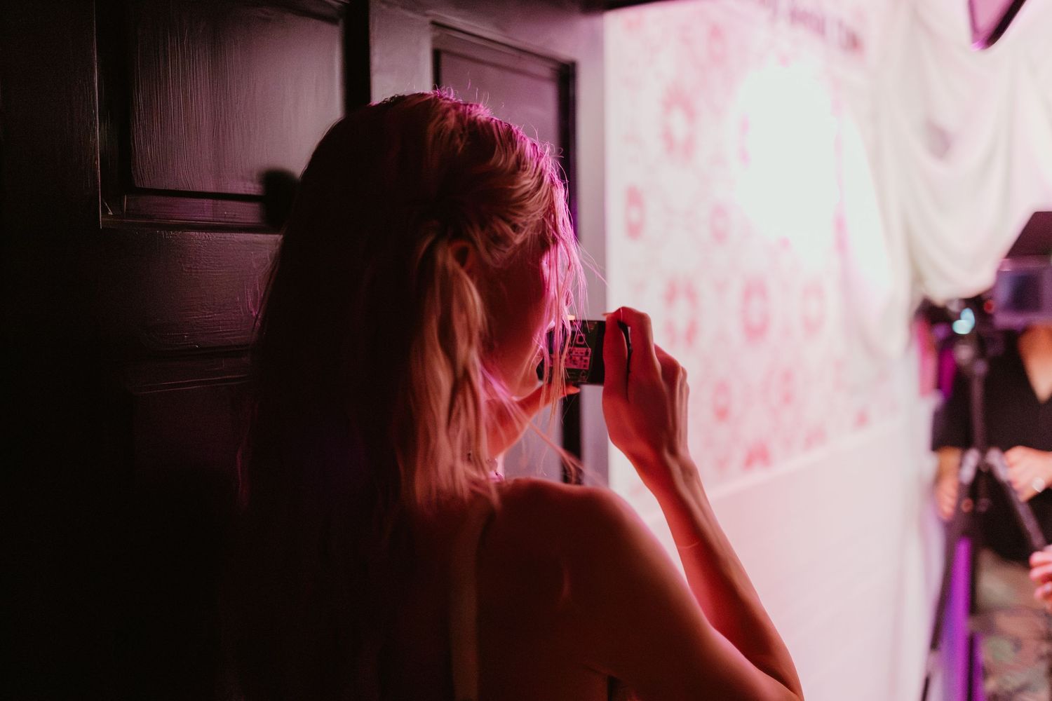 Woman with blonde hair photographing a decorated wall under pink lighting in a dimly lit room.