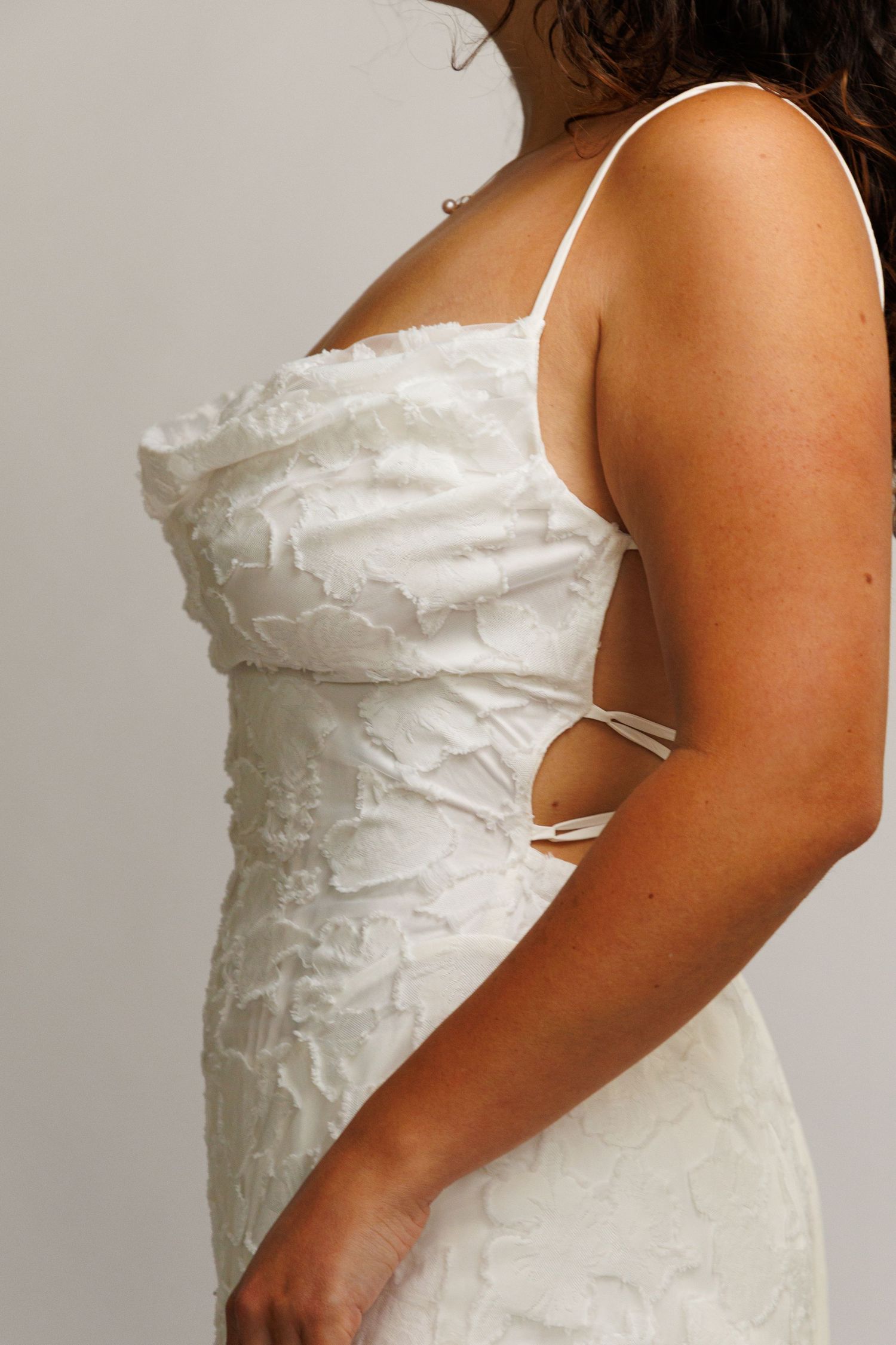 Woman wearing a white textured floral lace spaghetti strap bridal gown with open back tie detail.