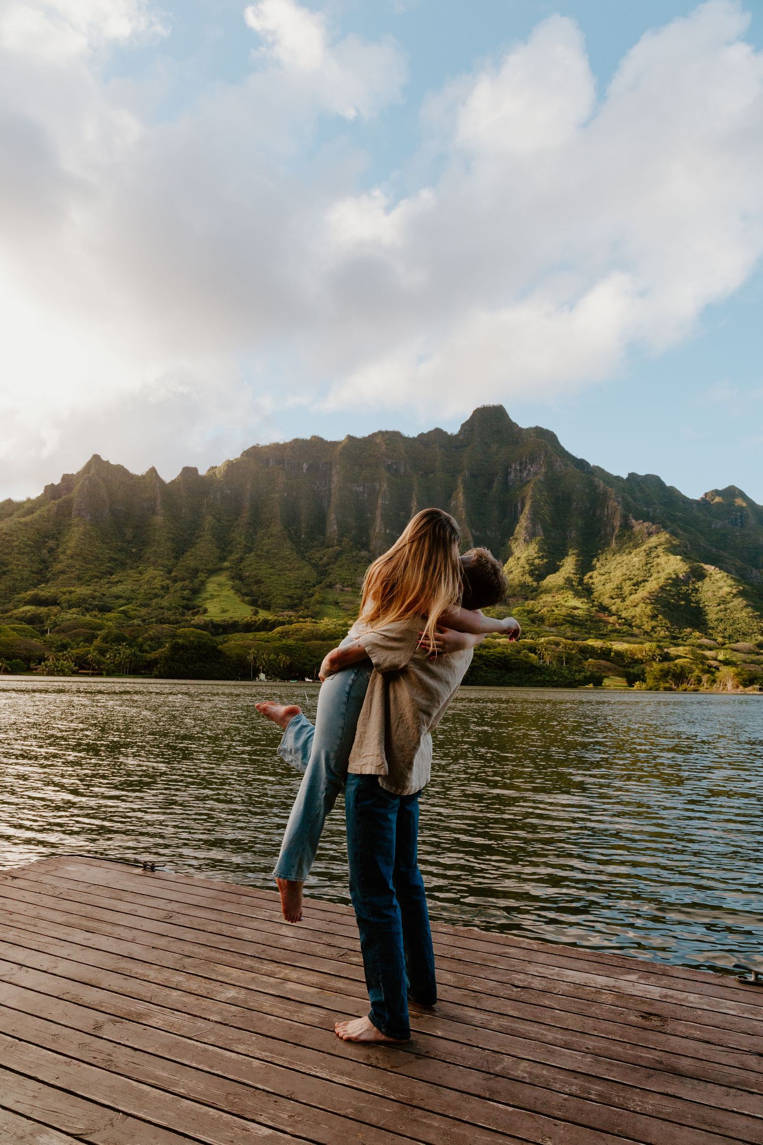 A couple embraces on a wooden dock with dramatic green mountains and a lake in the background.