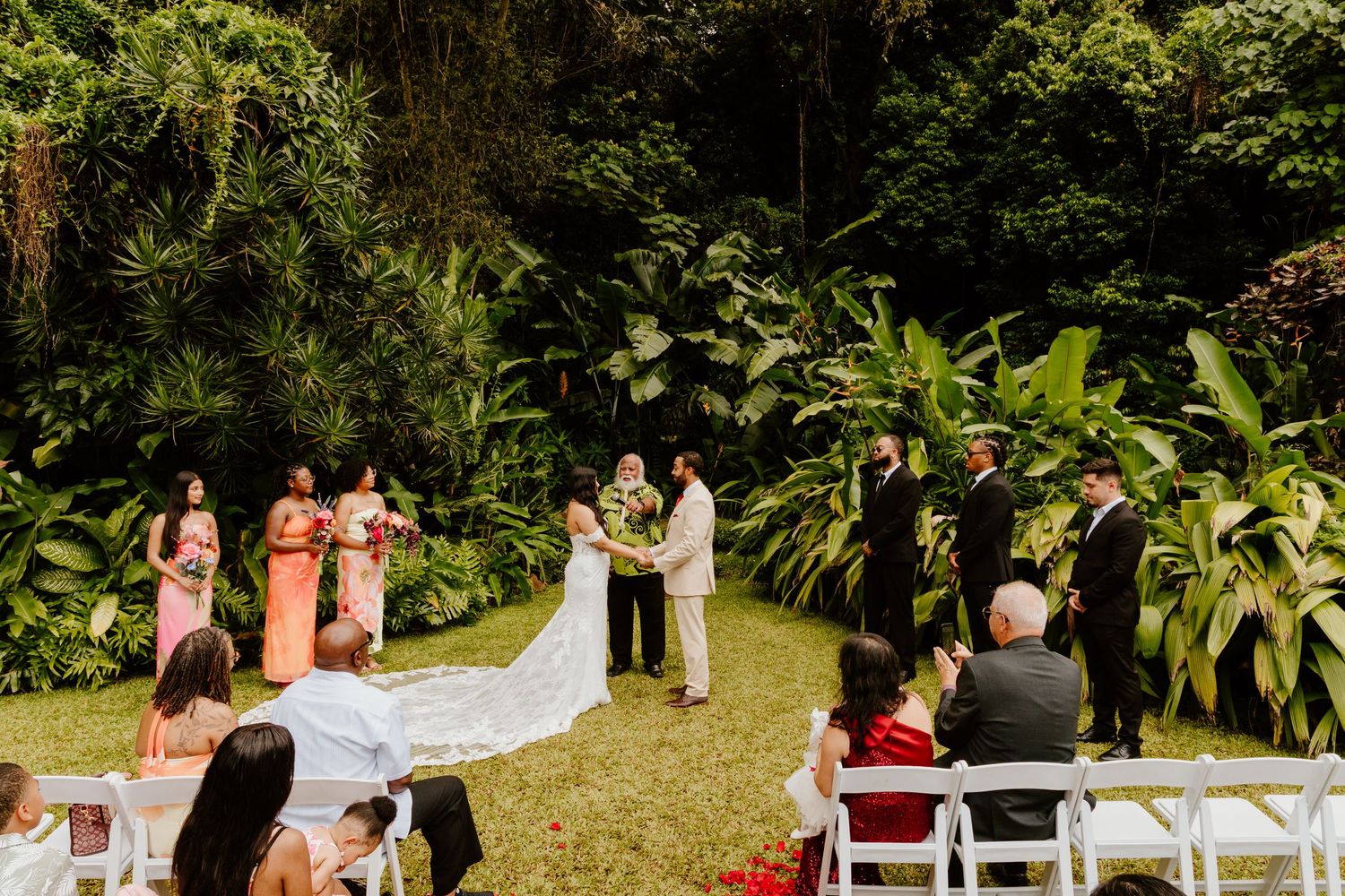 Outdoor wedding ceremony on lush green lawn with bridesmaids, groomsmen, and guests seated in white chairs amid tropical foliage.