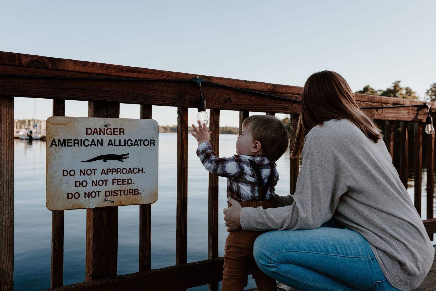 Mother and toddler crouch by a Danger American Alligator warning sign on a wooden lakeside dock at dusk.