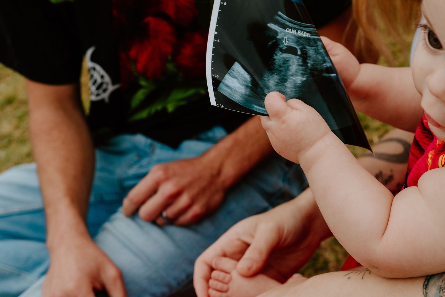 Baby reaching for ultrasound photo held by parent during outdoor family session.