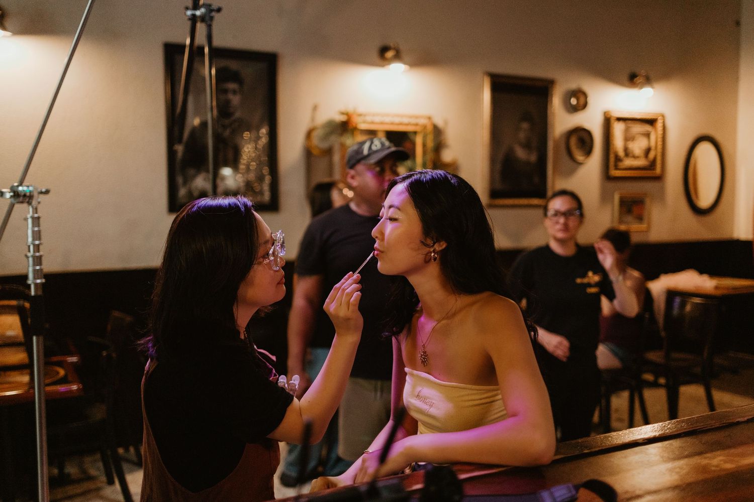 Makeup artist applying lipstick to a woman in a warmly lit restaurant setting with people in the background.