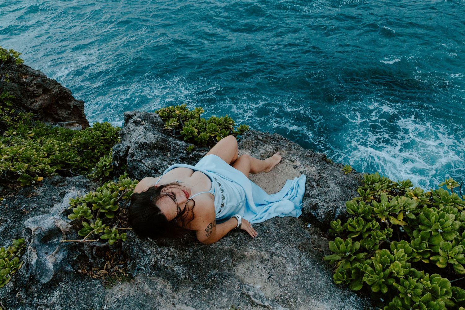 Woman in blue dress lying on rocky coastal cliff with turquoise ocean waves below.