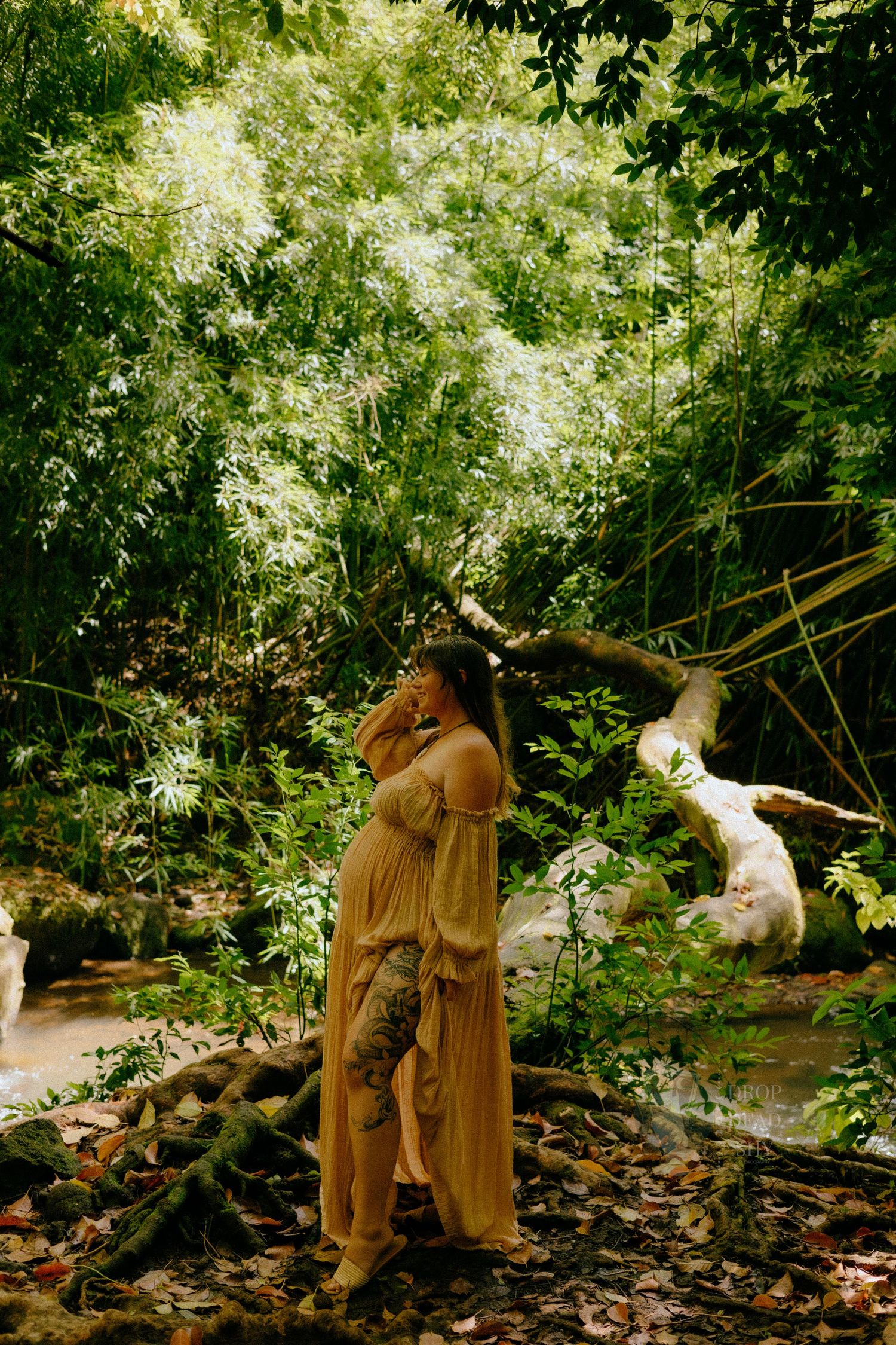 Pregnant woman in sheer peach dress standing near stream in tropical jungle with tattoos.