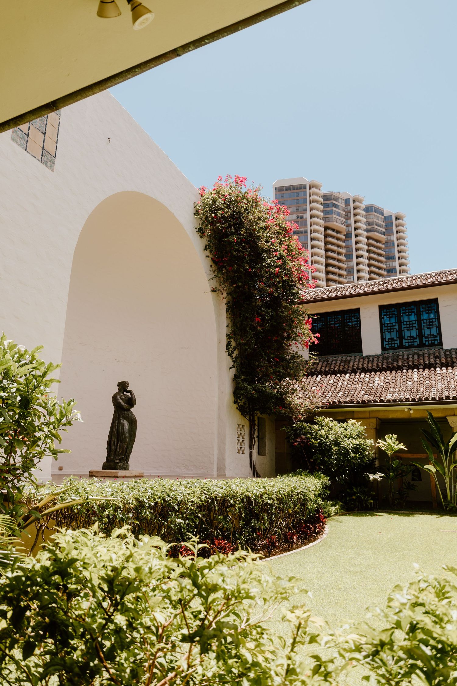 Courtyard garden with bronze statue, white arched wall covered in bougainvillea, and modern skyscraper in background.