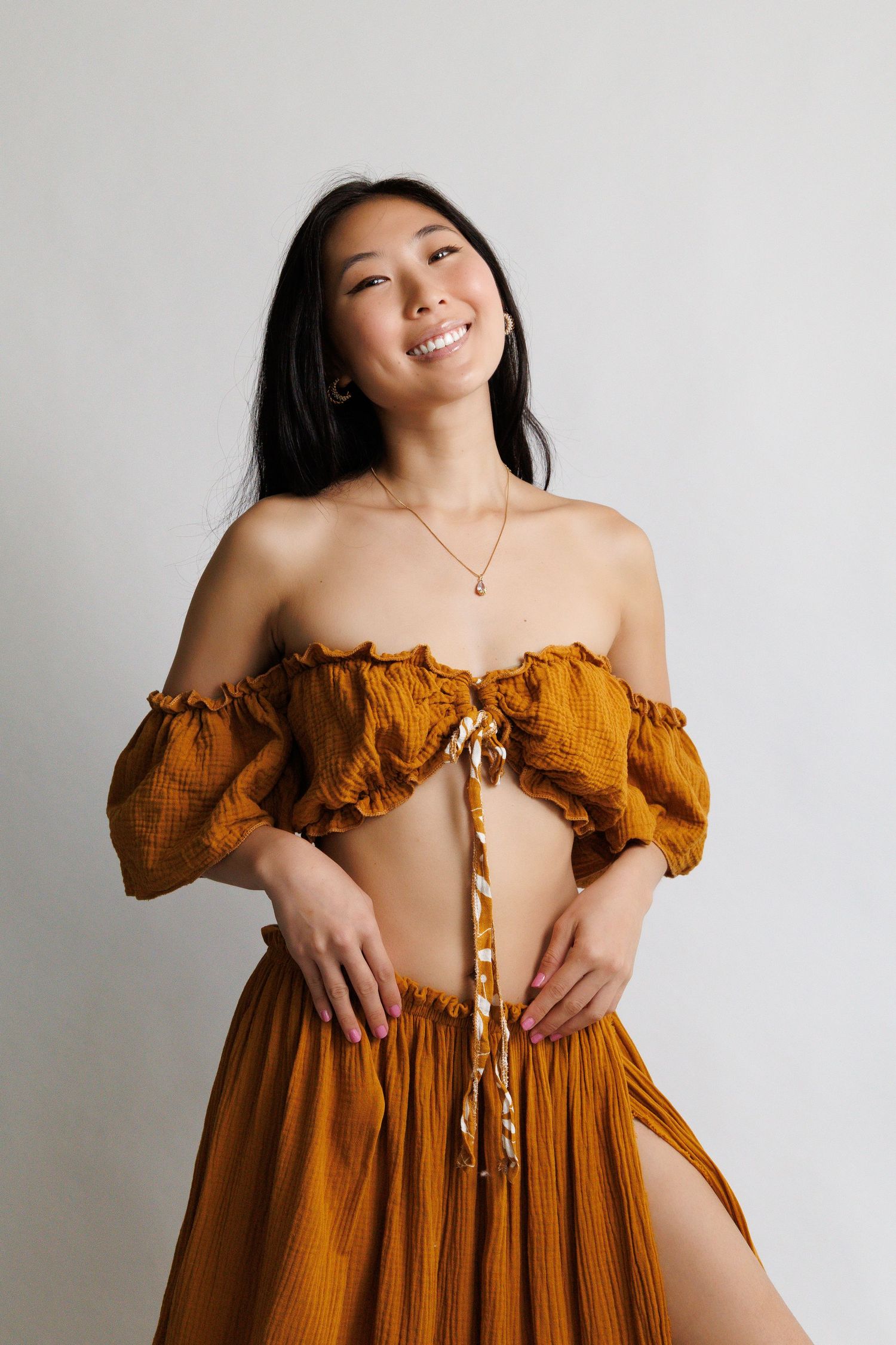 Woman smiling in mustard yellow off-shoulder crop top and matching maxi skirt with slit, posing against white background.