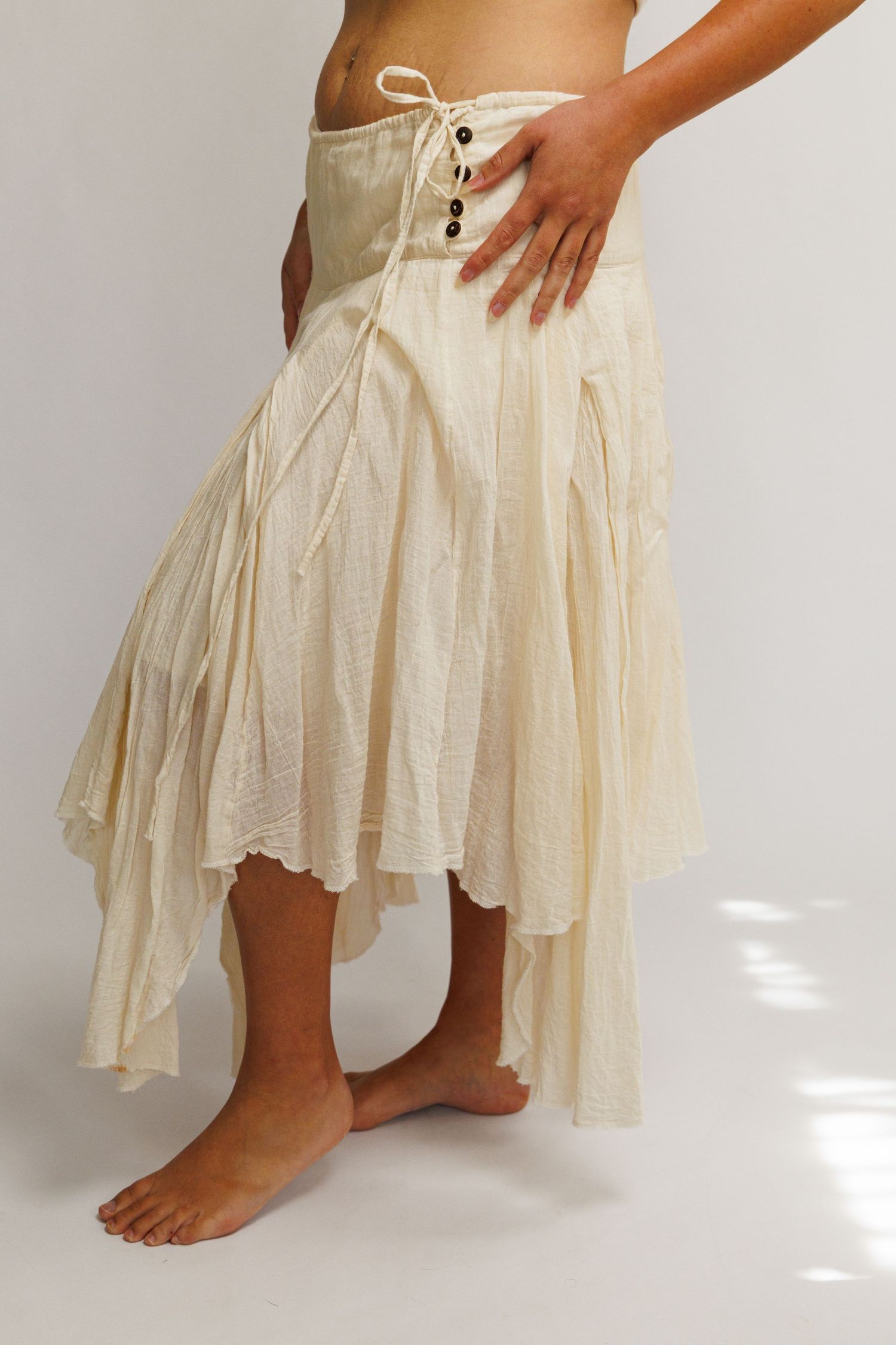 Cream bohemian asymmetrical skirt with lace-up waist detail, flowing layers, worn barefoot on light background.