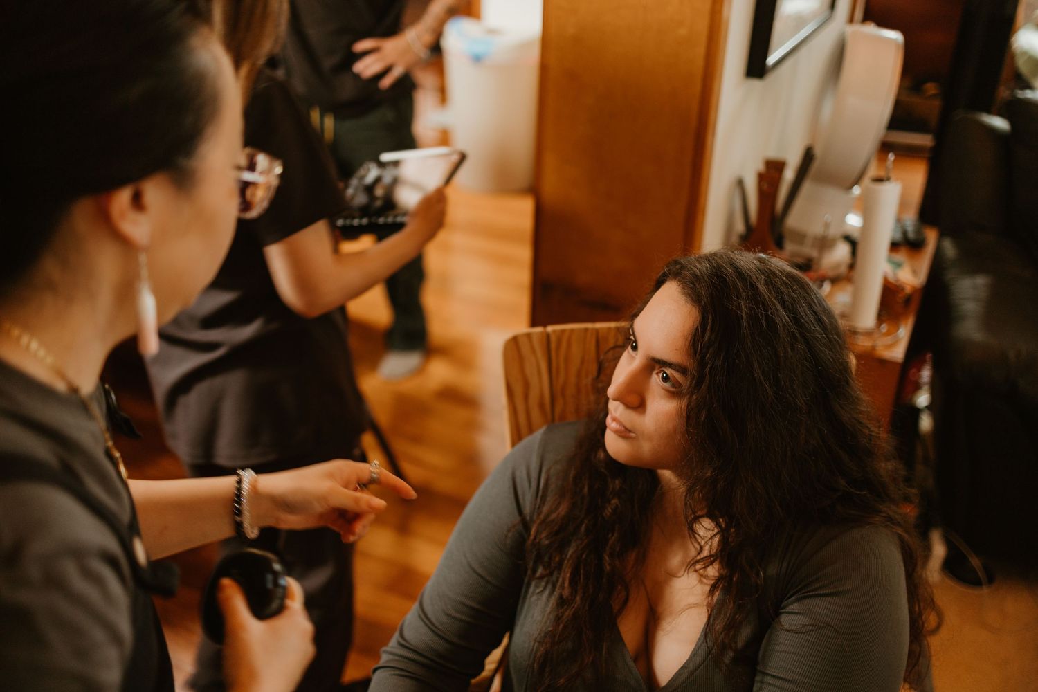 Woman with curly hair sits in chair while makeup artist works on her in a warmly lit studio.