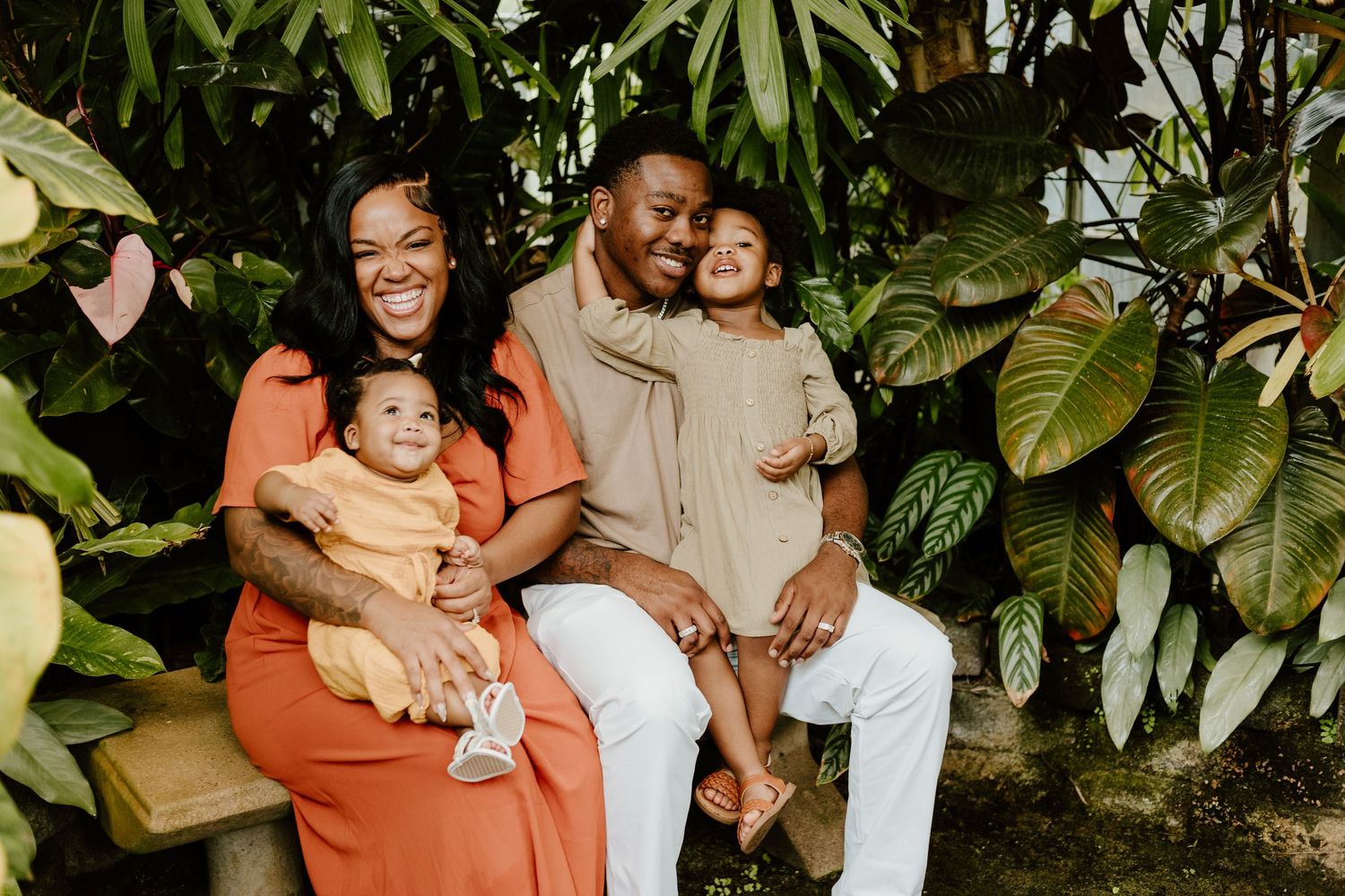 A happy family of four poses together on a bench surrounded by tropical greenery.