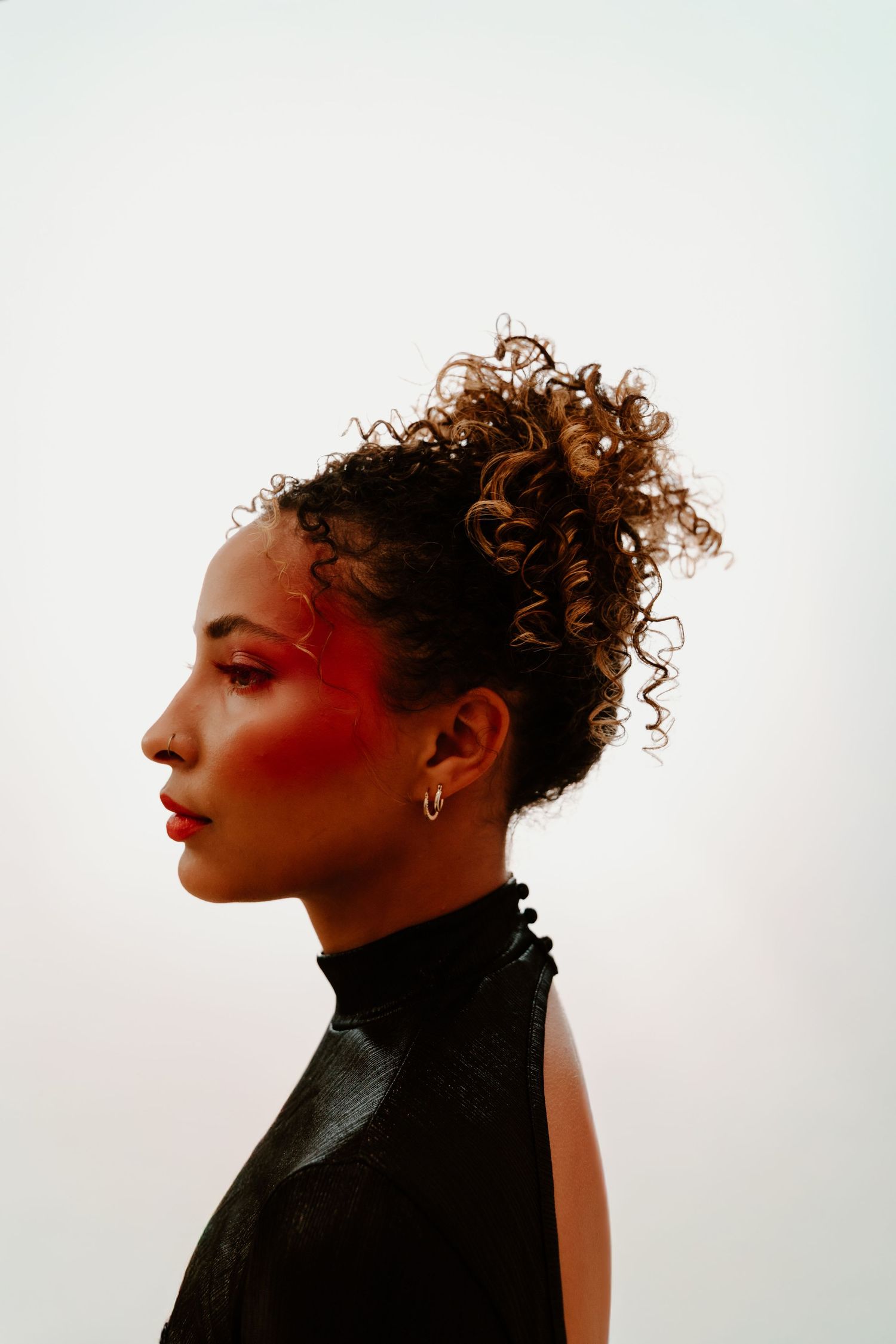 Profile portrait of a woman with curly updo hair, bold red eye makeup, and hoop earring wearing a black sleeveless turtleneck.