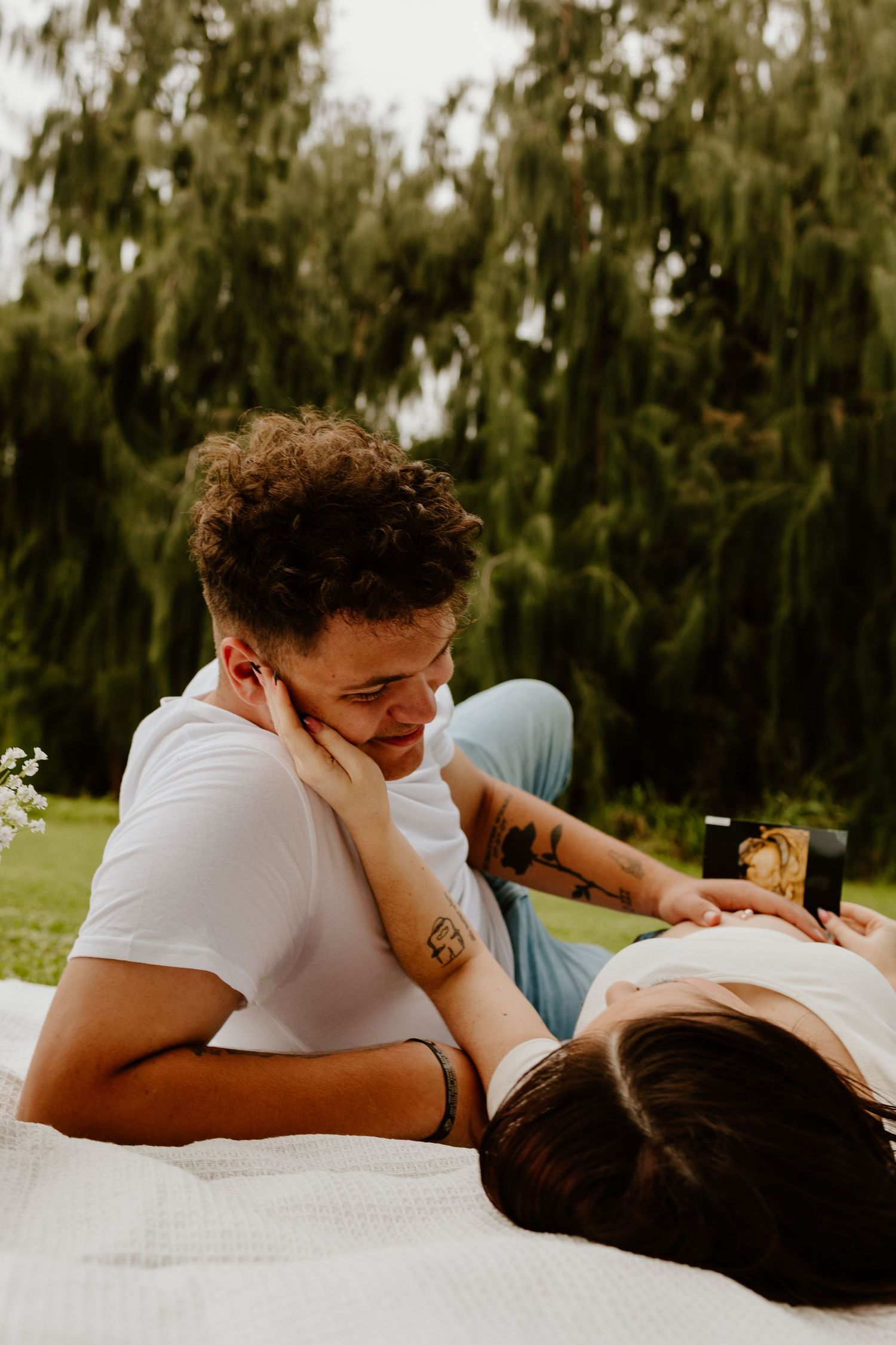 A curly-haired man gazes lovingly at a woman lying down on a white blanket in a park.
